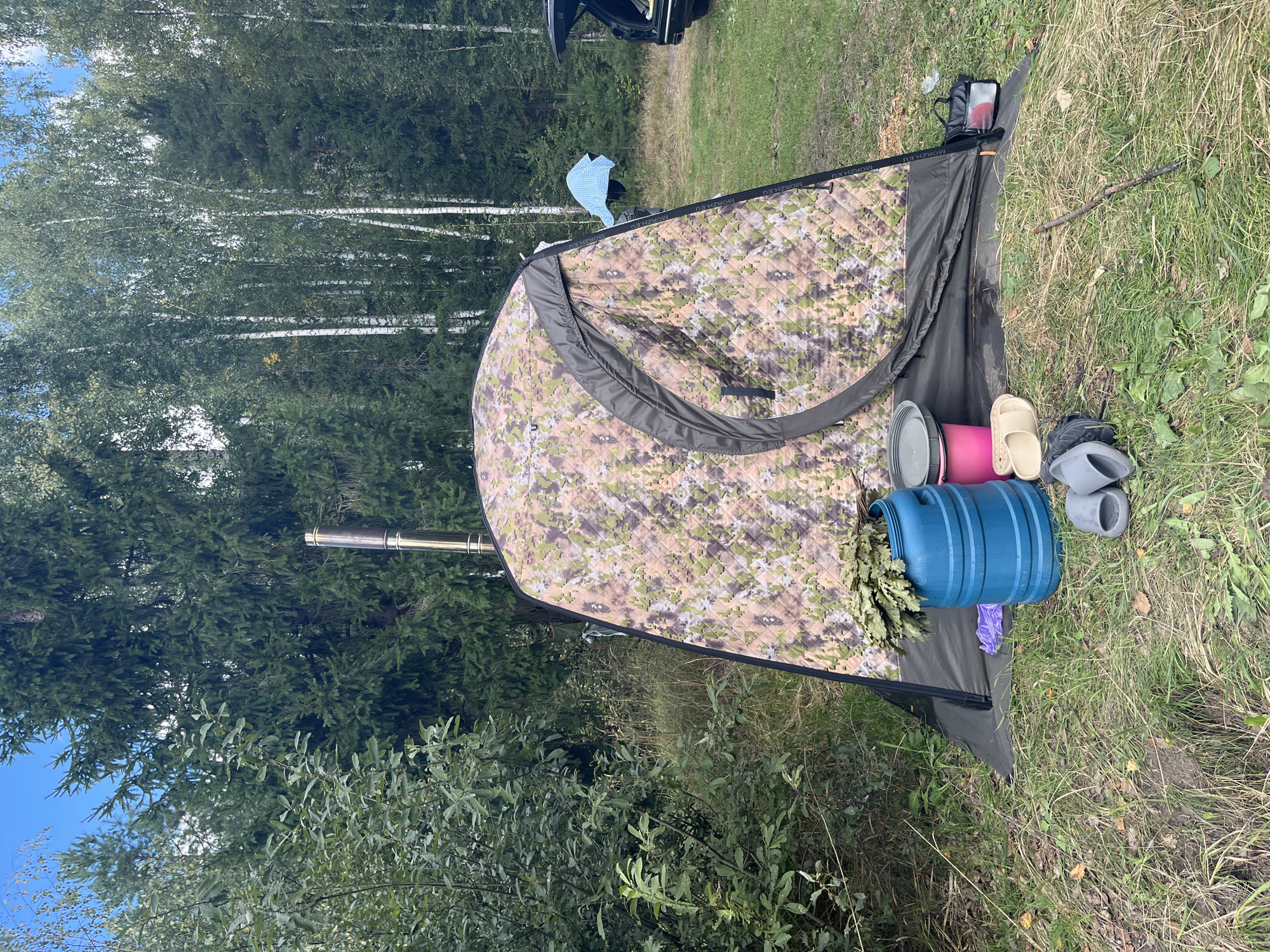 The image depicts a campsite in a forested area, likely in Novyi, Russia, given the presence of birch trees. In the foreground, a camouflage tent with a metal chimney is set up on a grassy clearing. Various items are placed around the tent, including a large blue barrel filled with birch branches (likely for a banya or sauna), a pink bucket, a grey lid, a pair of purple gloves, and two pairs of slippers. Behind the tent, a portion of a dark-colored SUV with its trunk open is visible, along with some laundry hanging on a line. The background is dominated by dense forest, with a mix of evergreen and deciduous trees under a clear blue sky. The overall scene suggests an outdoor recreational activity, possibly involving a banya experience in nature.