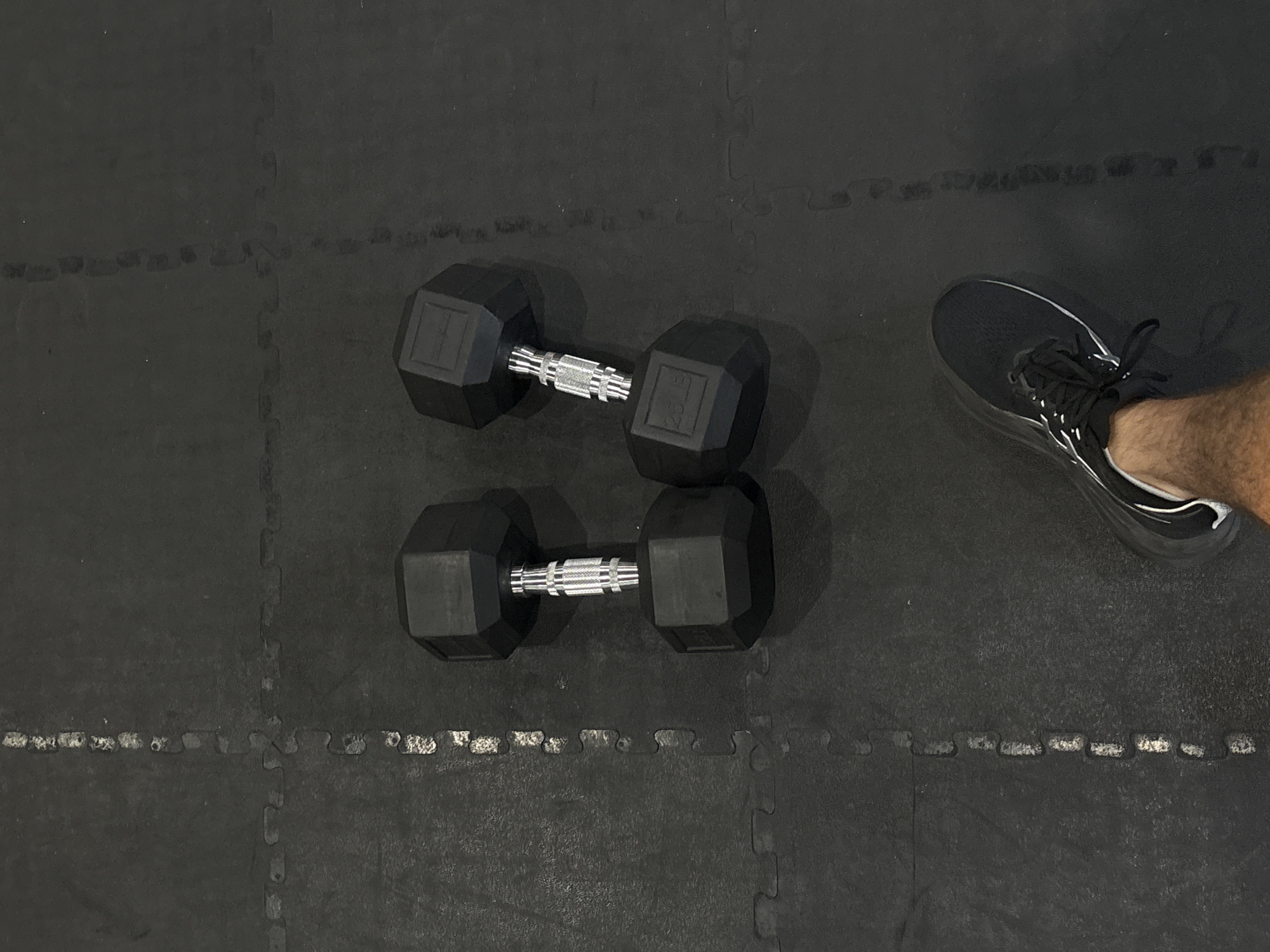 A pair of black hexagonal dumbbells, each appearing to be 25 lbs, are arranged vertically on a black interlocking gym floor. A foot clad in a black running shoe is visible in the upper right corner of the frame. The location is assumed to be in San Bernardino Tlaxcalancingo, Mexico, but there is no forensic evidence within the image to confirm this.