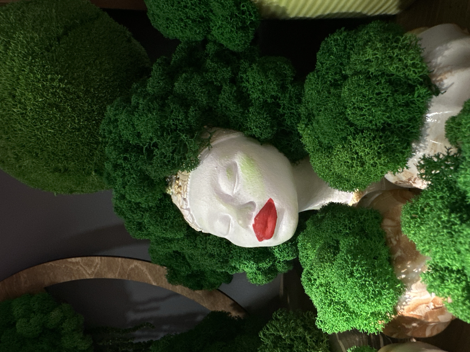 A white, featureless mannequin head with closed eyes, prominent red lips, and a golden-cream decorative band on its forehead is centrally displayed. The head is adorned with vibrant green, preserved moss or lichen, forming an elaborate, voluminous hairstyle. Surrounding the mannequin are numerous clusters of the same bright green, textured material, some appearing as large spherical formations and others as smaller, irregular mounds. These green elements rest on various light-colored, possibly ceramic or stone, bases. The background is dark and muted, with a segment of a curved, light brown wooden structure visible on the upper left. This decorative display is located in Rostov-on-Don, Russia.