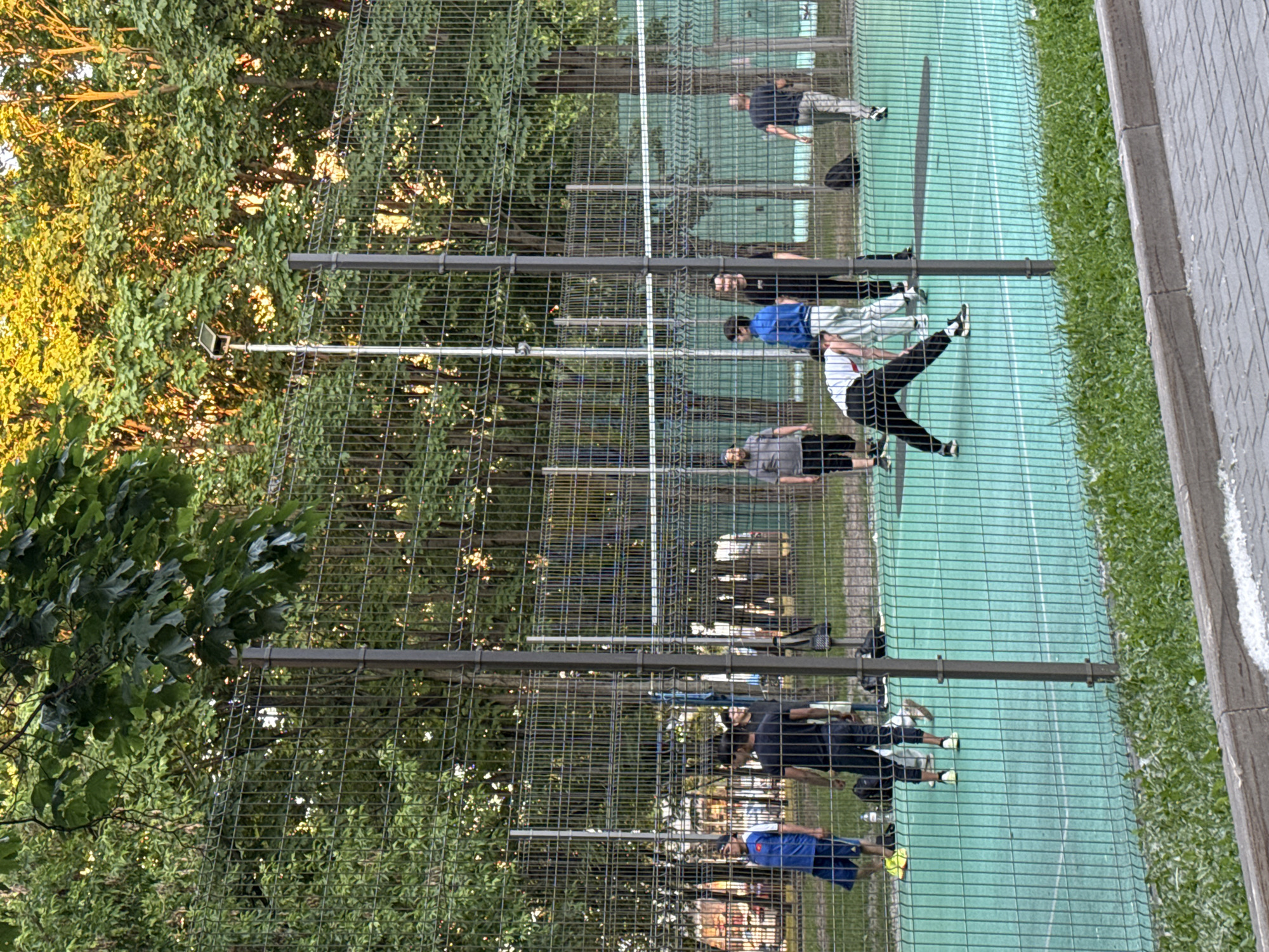 The image depicts an outdoor sports court, likely for volleyball, in Moscow, Russia, during late afternoon or early evening under warm, golden sunlight filtering through trees. The court features a teal-green playing surface with white boundary lines and is enclosed by a dark grey wire mesh fence supported by robust metal posts. A white net is stretched across the court in the background.
Nine individuals, primarily adult males, are visible on or around the court, dressed in casual athletic wear. In the foreground on the court, one person in a white top and black pants is bent at the waist, appearing to stretch or reach for something. To their left, another individual in a grey t-shirt and dark shorts stands. Further left, a person in a blue t-shirt with a red emblem and dark shorts stands facing right. In the middle ground, near the net, two individuals in dark t-shirts are standing. On the far left beyond the court, another person is partially visible. On the far right, a person in a grey top and light pants walks away from the court. Another person in a dark top stands behind the stretching individual.
Various personal items, including dark bags and water bottles, are placed on the court's perimeter. A tall, thin metal pole with a small, rectangular apparatus (possibly a light fixture or camera) is positioned behind the net. The court is surrounded by numerous deciduous trees with dense green foliage, with sunlight illuminating their upper branches. In the immediate foreground, a concrete curb separates a strip of green grass and weeds from a paved area of interlocking grey pavers.
