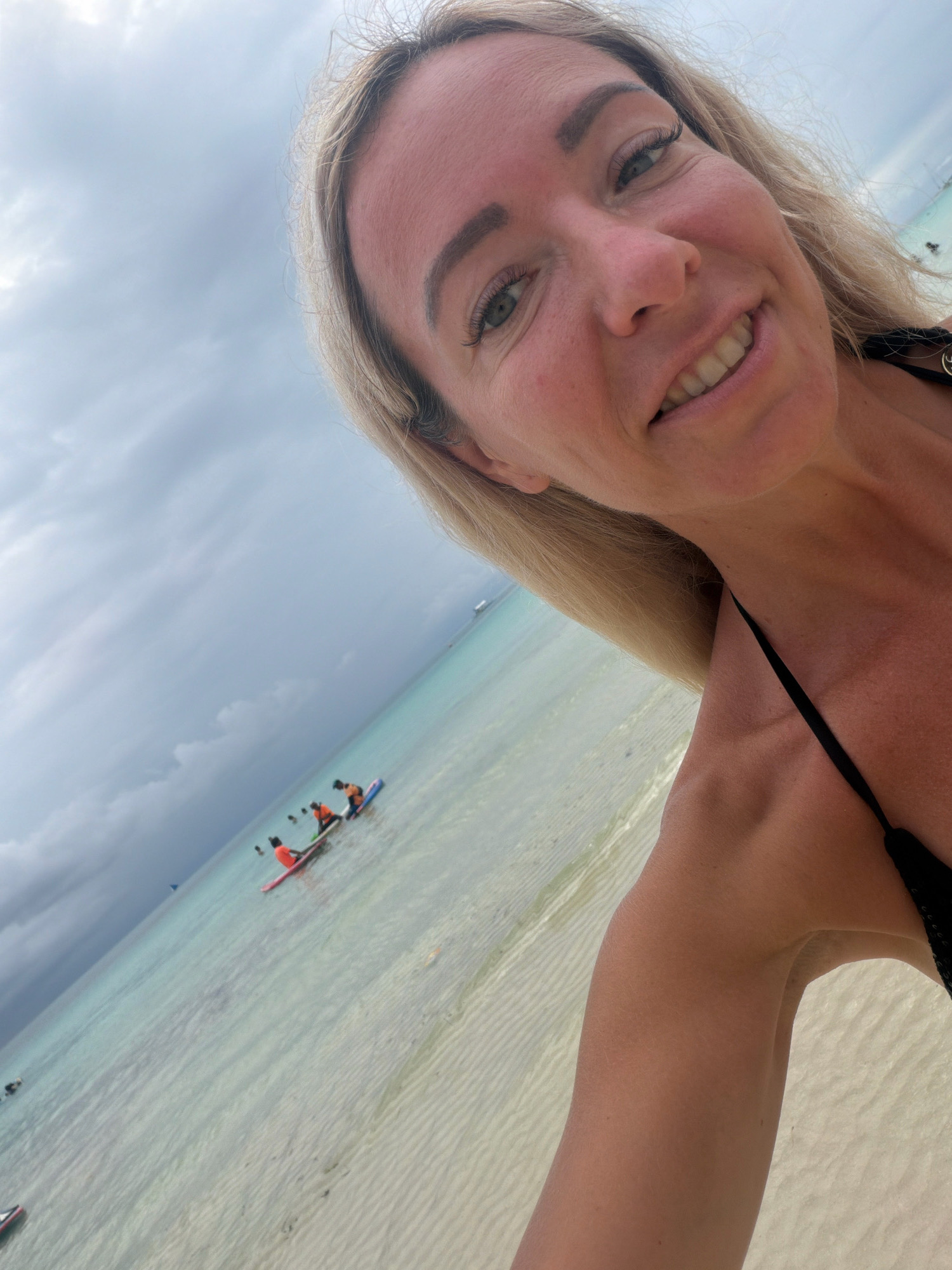 A woman with blonde hair and green eyes is taking a selfie at a beach. She is wearing a black bikini. In the background, there are several people on paddleboards in the ocean. The water is clear and shallow, and there is a sandy beach. The sky is cloudy. The location is likely a beach in Malay, Philippines, possibly Boracay Island given the activity of paddleboarding in shallow water and the overall scenery.