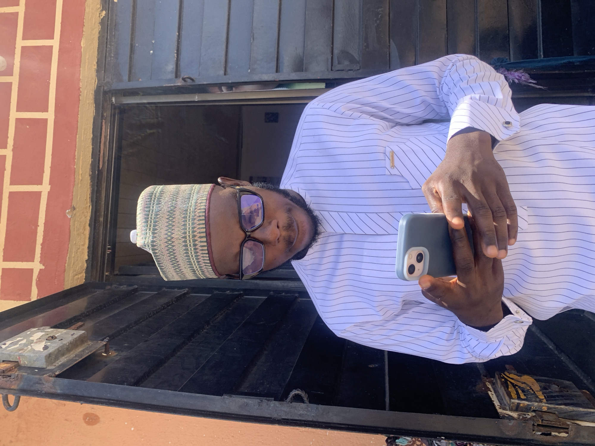 A person wearing a white striped shirt, a patterned hat, and glasses is holding a blue smartphone and looking at the camera. The person is standing outside a building. Part of a brick wall is visible, as well as a dark metal structure, possibly a window covering or gate. The scene appears to be outdoors, with ample natural light. It is unknown if the location is indeed Jos, Nigeria.