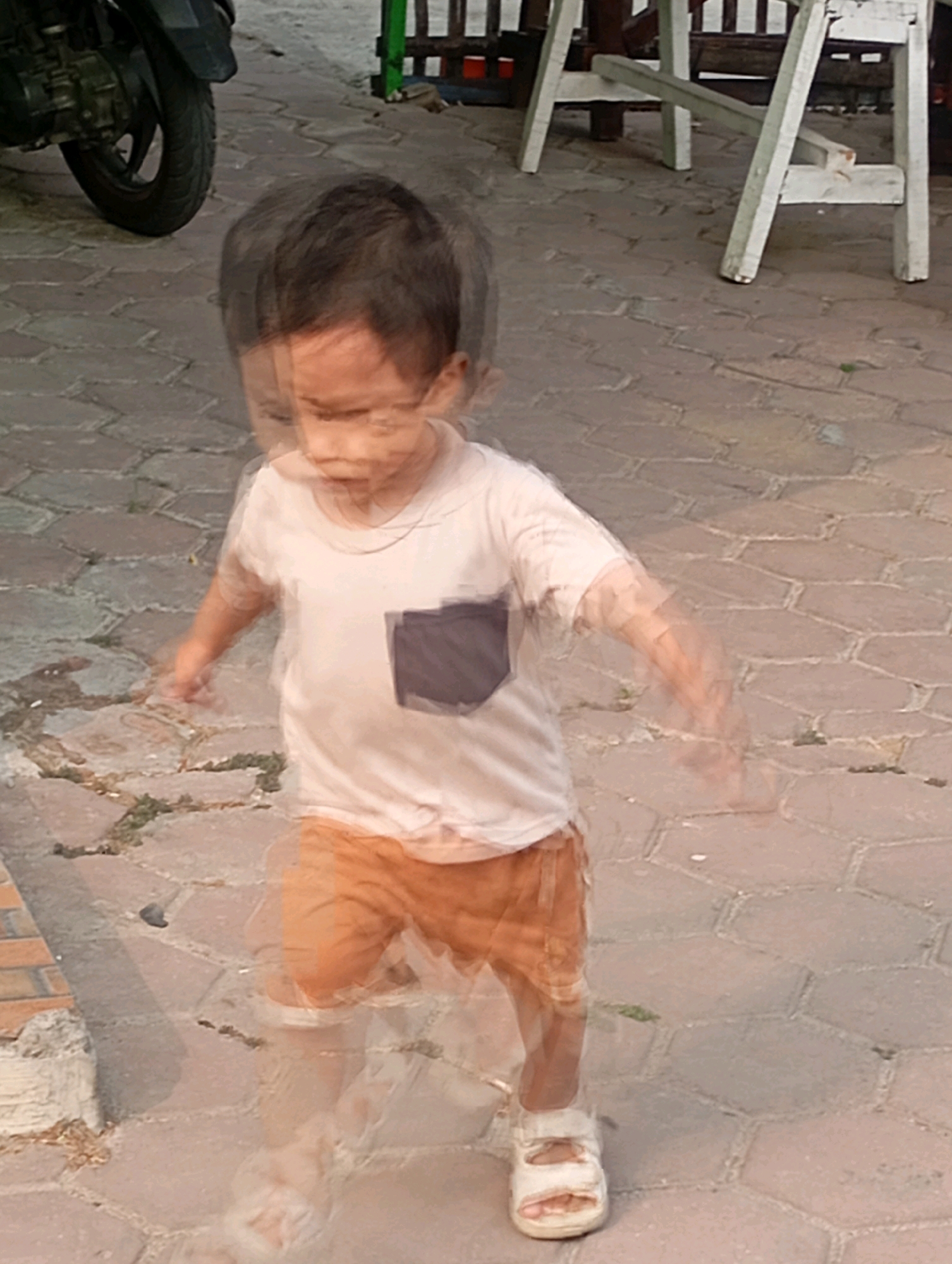 The image shows a child in motion, captured with a motion blur effect, on a paved outdoor area. The child, appearing to be a toddler, is wearing a light-colored t-shirt with a dark rectangular patch on the chest and orange shorts. They are also wearing white sandals. The ground is paved with hexagonal bricks of varying shades of gray and brown. To the left of the child, the rear tire and part of a motorcycle are visible, suggesting an urban or semi-urban setting. In the background, a white wooden structure, possibly part of a table or seating area, is visible, along with some greenery.
The scene is located on Jalan Bahagia Raya, Babelan, Indonesia. This street is a common thoroughfare in the area, characterized by residential and commercial establishments, as well as public spaces. The presence of the motorcycle and the paved ground are consistent with the typical urban landscape of Babelan. The motion blur of the child indicates activity, possibly playing or walking. The overall impression is of an everyday scene in a public area of Babelan.