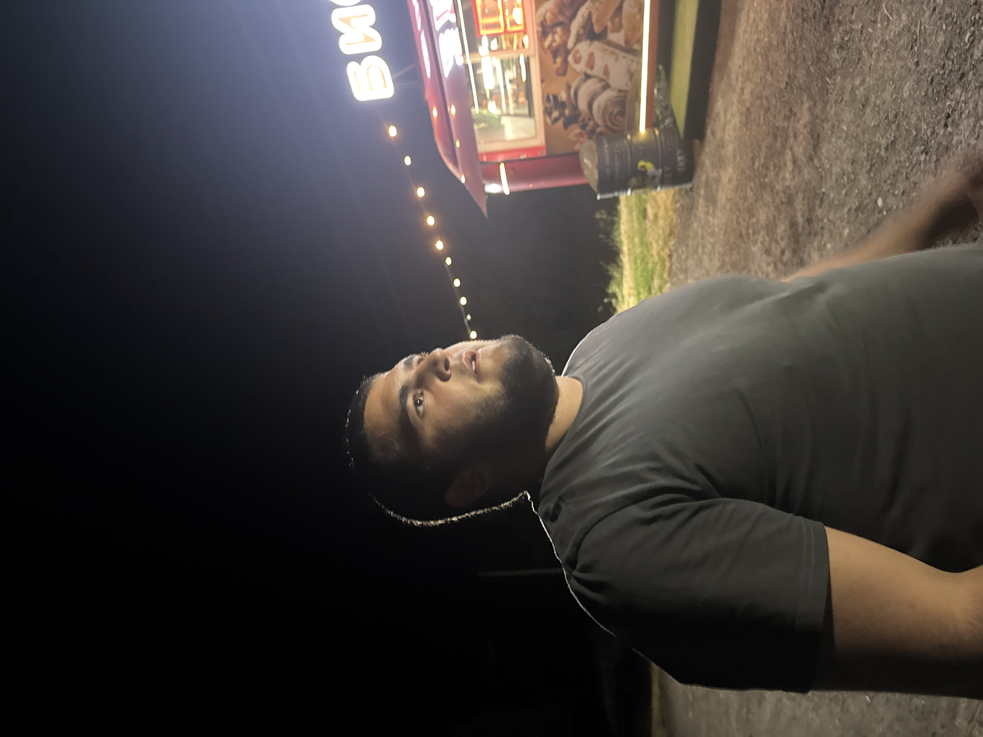 The media file captures a night scene featuring a man and a food truck in Yutin, Russia. The man, positioned in the foreground, is looking upwards and to his left. He has dark hair and a beard. He is wearing a dark grey t-shirt.
The food truck, a prominent object in the background, is illuminated and appears to be open for business. It has a red exterior with yellow lettering. The visible text on the truck appears to be in Cyrillic script, with "ВЫПЕЧКА" (Vypechka, meaning "Baking") clearly legible. Images of food, possibly pastries or baked goods, are displayed on the side of the truck. A string of lights hangs above and to the left of the truck, casting a warm glow. Next to the truck, a dark cylindrical object, possibly a barrel, is visible.
The ground surrounding the food truck and man is a gravelly, dusty surface. Some dry grass and vegetation are present to the right of the food truck. The overall atmosphere is that of an outdoor evening gathering or a street food vendor operating at night. The lighting suggests it is dark outside, likely evening or night. There are no clear indications of weather conditions, but the scene appears dry.