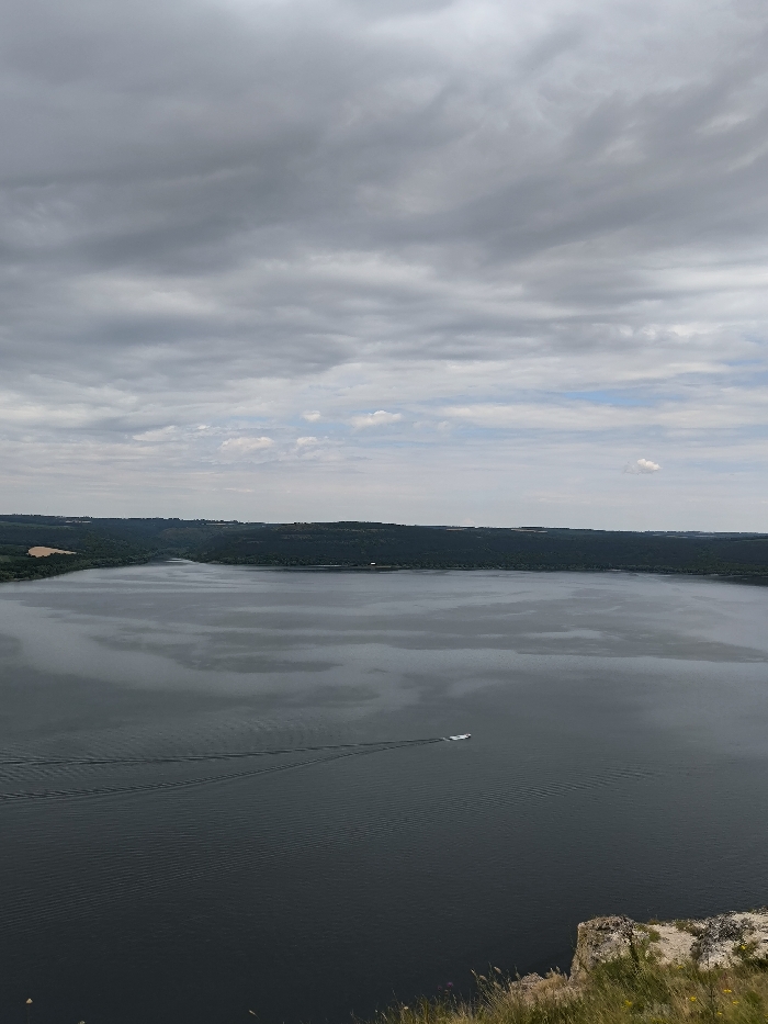 The image presents a wide panoramic view of a large body of water, identified as a section of the Dniester River or Dniester Reservoir, observed from an elevated cliffside vantage point in Horaivka, Ukraine. The upper two-thirds of the scene are occupied by a predominantly overcast sky, characterized by extensive grey stratiform clouds with minimal blue sky visible.
The central and lower portions of the image feature the expansive, dark grey water surface, displaying subtle ripples. A single white motorboat is positioned on the water, slightly right of the vertical center, moving from right to left across the frame. It leaves a distinct, bifurcated wake extending behind it towards the lower-left. The far bank of the water body, visible along the horizon, consists of a continuous, dark green, heavily forested landmass.
In the immediate foreground, at the bottom right, a segment of a light-colored, weathered rocky cliff face is present, with sparse green grasses and yellow flora growing along its edge. No individuals are visible within the scene. The overall lighting is diffused, consistent with the cloudy conditions.