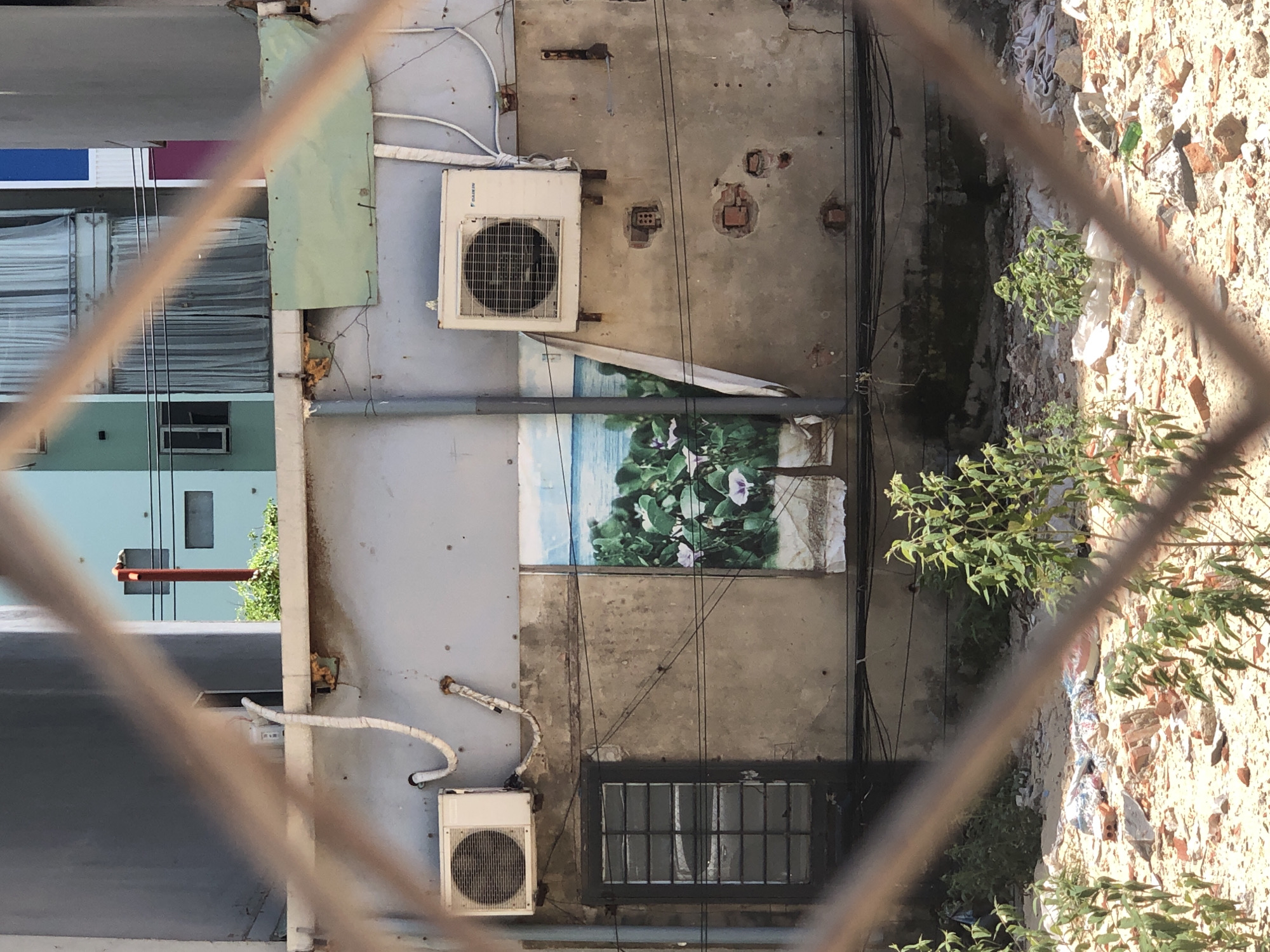 The image shows a gritty, urban scene viewed through a diamond-patterned fence. In the foreground, the fence bars are out of focus, with some vegetation growing below. Behind the fence, a weathered concrete wall dominates the midground. Two white air conditioning units are mounted on this wall, one on the left and another on the right. Between them, a faded mural depicting a tropical scene with green foliage and purple flowers against a blue sky and water is affixed to the wall. To the left of the mural and below the left air conditioning unit, a dark-colored door with a grid of windows is visible. The wall itself is scarred with exposed bricks and has numerous wires running across it. In the background, a lighter-colored building with windows and draped curtains can be seen. The ground in front of the wall is covered with debris, including bricks, sand, and discarded items. The overall impression is one of urban decay and neglect.