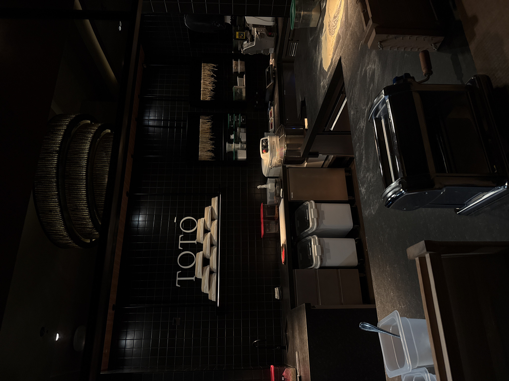 The image depicts the interior of a pasta-making kitchen, specifically the Toto restaurant in Dubai, United Arab Emirates. The scene is dimly lit, with a focus on the work area. The kitchen features a black tiled wall with the name "TOTO" displayed vertically in white. Near the wall are stacks of what appear to be pasta sheets or ingredients. A metal pasta machine is prominently placed on a countertop, suggesting active food preparation. Other kitchen equipment and storage containers are visible.