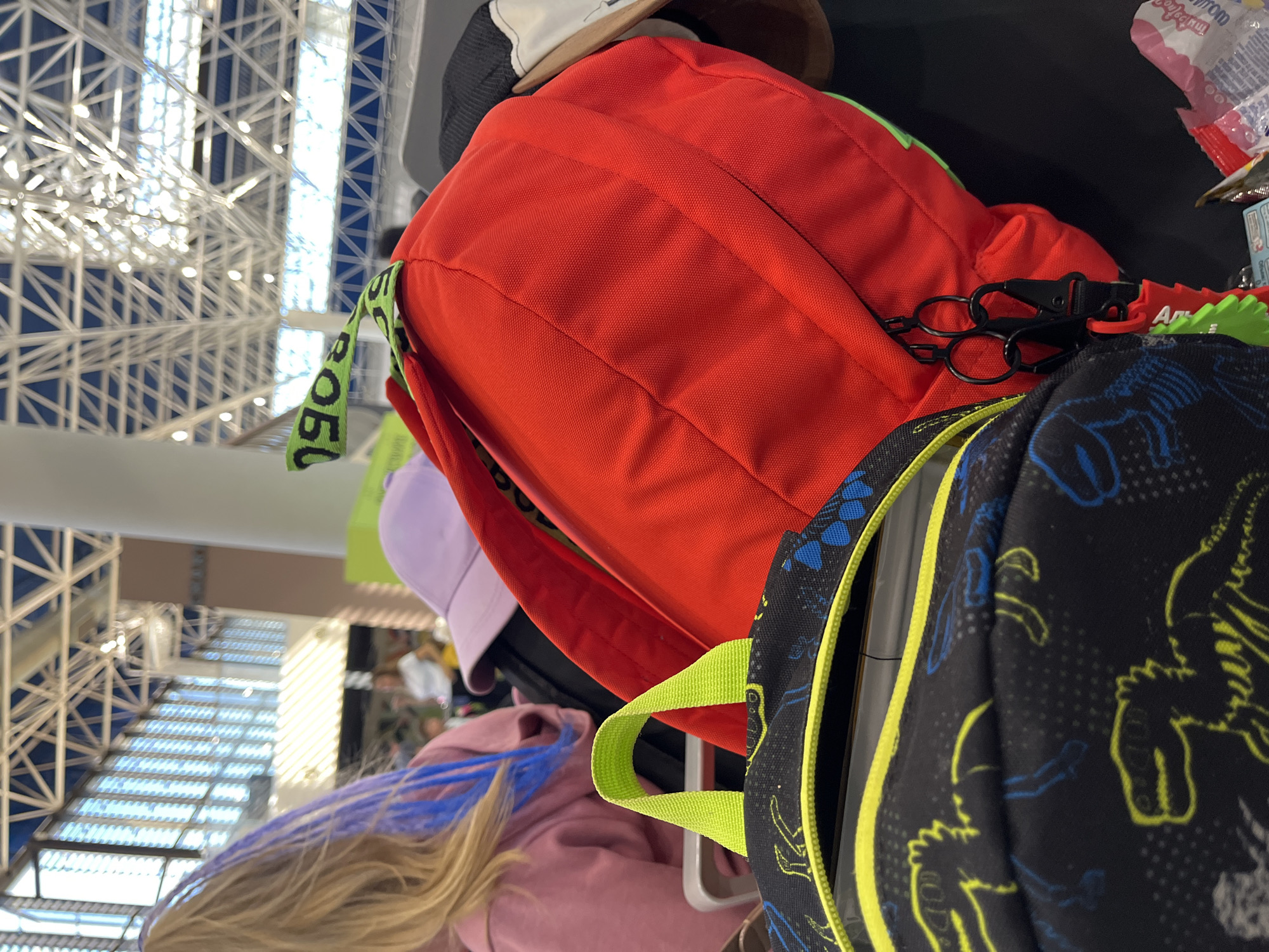 The image shows a close-up view of several backpacks and a purple baseball cap, likely in an indoor waiting area or public space. In the foreground, a black backpack with a dinosaur pattern and a bright yellow zipper is visible. Behind it, a red backpack with a green tag and black zipper pulls is prominent. To the left, the blonde hair of a person with blue streaks is visible, wearing a pink hoodie. A purple baseball cap rests on top of another object. In the background, a person in a white shirt is partially visible. The ceiling structure above appears to be a modern architectural design, possibly with skylights, suggesting a large public building such as an airport or a train station. The overall lighting indicates an indoor environment with diffused natural light. The image does not contain any specific landmarks or clues that would definitively place it in Sochi, Russia.