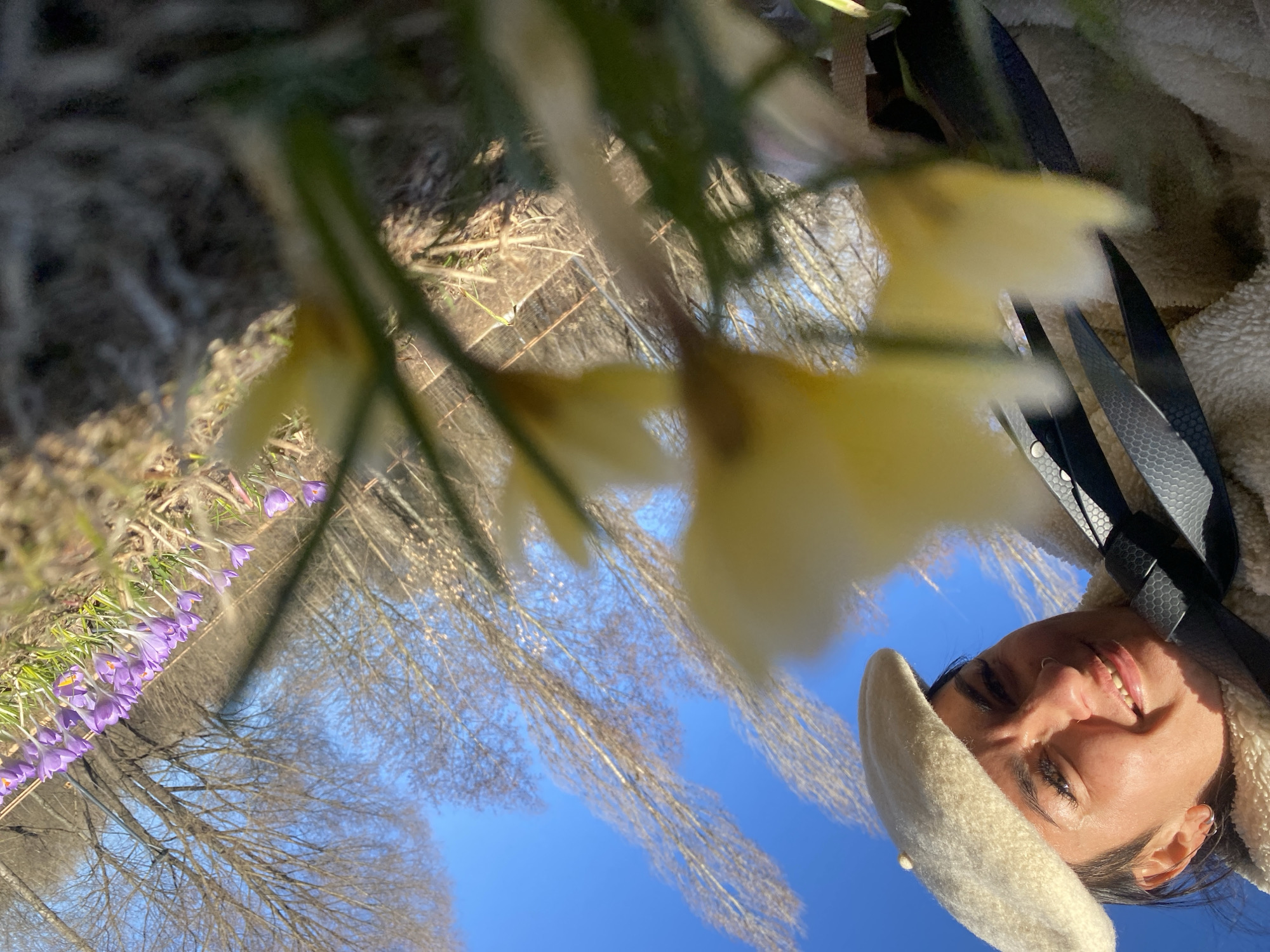 The photograph depicts a woman, wearing a cream-colored beret and a cream-colored sherpa-style jacket, smiling at the camera. Black straps are visible near her shoulder. The image is taken from a low angle, with out-of-focus yellow flowers in the foreground. A row of purple crocuses is visible on the left side. The background includes bare trees and a bright blue sky. Without additional information, the location in Moscow, Russia cannot be determined.