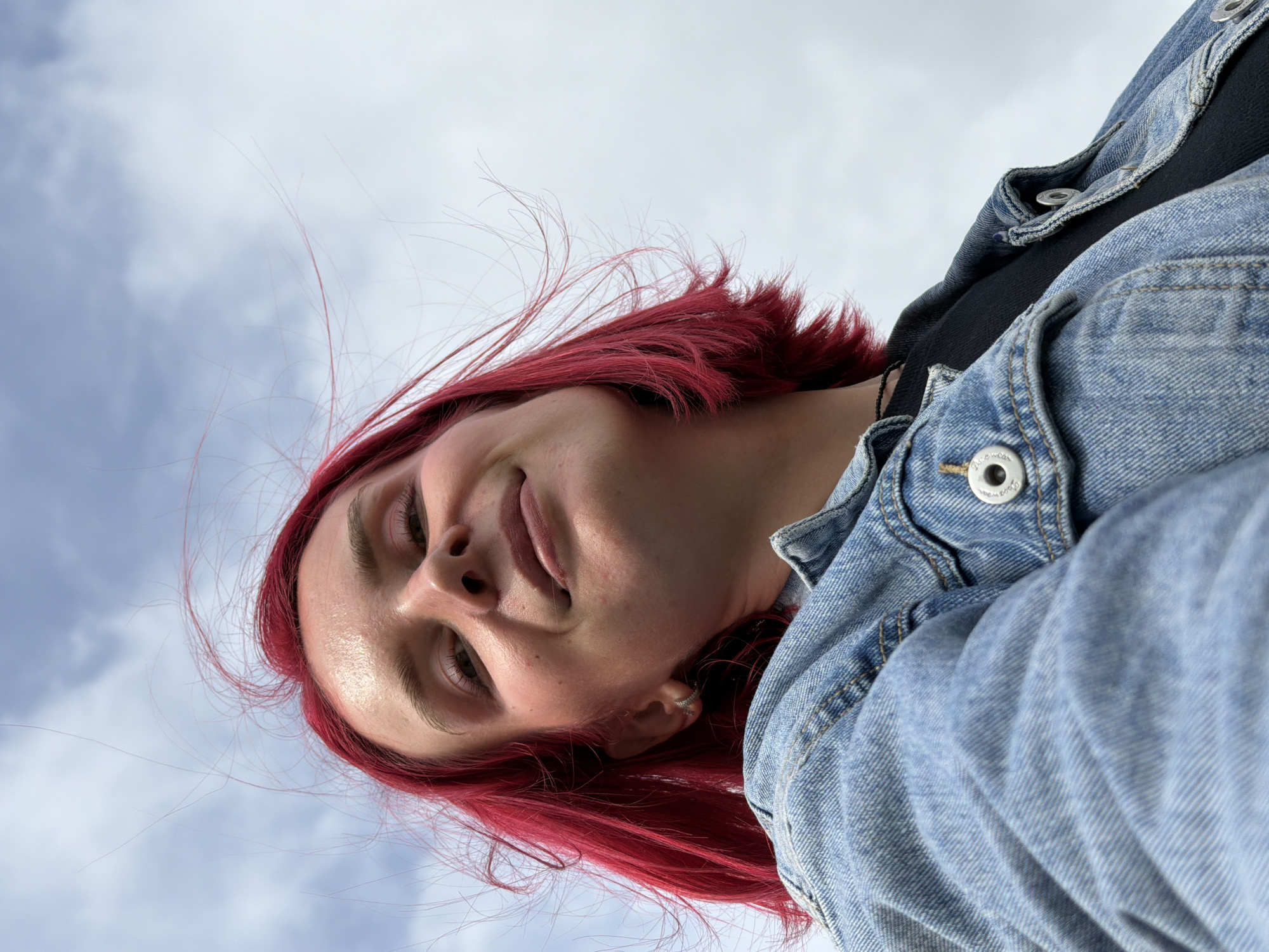 A low-angle portrait captures a young individual with vibrant red hair looking downwards into the camera. The subject has a subtle, relaxed smile or smirk and engaging eyes. Their skin appears dewy under the bright, natural light. They are dressed in a light blue denim jacket, with metallic buttons clearly visible, one of which displays the text "AMERICAN EXCHANGE." A dark-colored garment is worn beneath the jacket. A small silver hoop earring is visible in their right ear.
The background is an overcast or partly cloudy sky, indicating an outdoor setting during the daytime. Several strands of the person's bright red hair are wind-blown, suggesting a light breeze. The unique perspective looks up at the subject, making them appear dominant against the sky. The image portrays a casual, perhaps playful, moment in Chaikovsky, Russia.