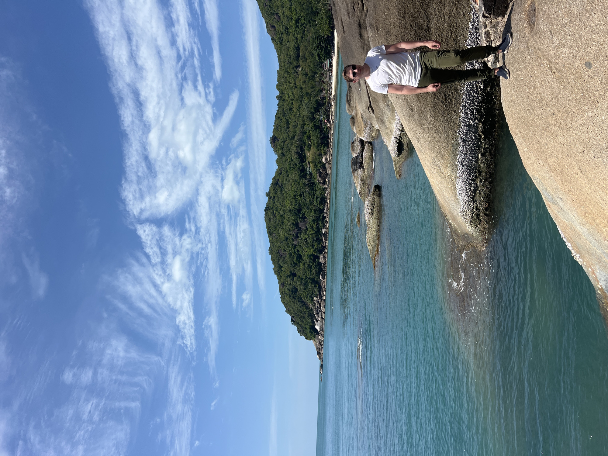 A man stands on a large rock formation overlooking a turquoise bay in Ko Samui District, Thailand. The man, wearing a white t-shirt and olive green pants, is positioned on the right side of the frame, facing forward with sunglasses on. The bay is calm with clear water, and several large rocks are visible both in the water and on the shore. In the background, a lush green hillside slopes down to a sandy beach with a few buildings. The sky is a bright blue with scattered white, wispy clouds. The scene appears to be a sunny day at a tropical beach.