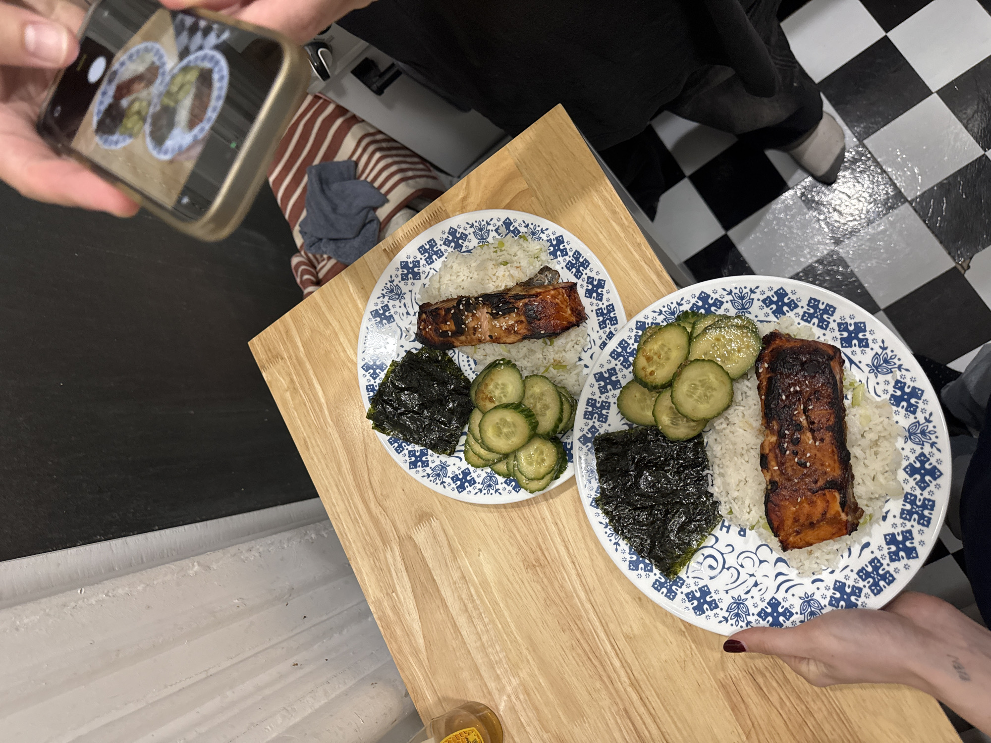 Two plates of food are on a wooden table, with a person holding up a smartphone to photograph them. The plates contain rice, seaweed, cucumber slices, and a rectangular piece of cooked protein. The food is presented on blue and white patterned plates. A person is taking the picture, holding the phone in their left hand, while another person holds the plate in their right hand. The background includes a checkered floor, striped fabric, and other pieces of furniture. The scene appears to be indoors, possibly in a kitchen or dining area. It is impossible to identify the location as New York City, or any city without further information.