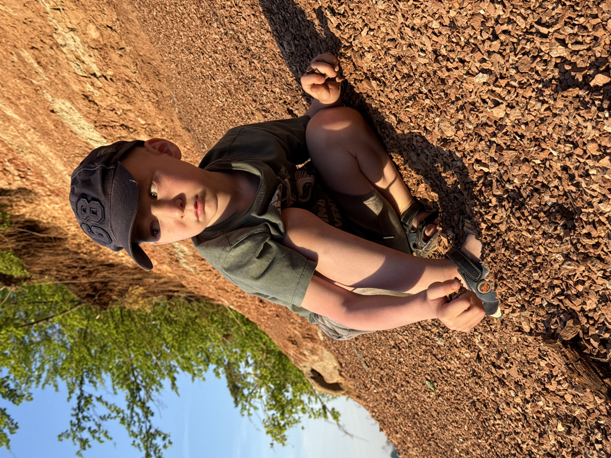 A young boy wearing a dark green t-shirt, gray shorts, sandals, and a dark blue baseball cap with a number 88 on it is sitting on a ground covered in reddish-brown gravel. He is holding a piece of gravel in his hand. Part of a tree is visible in the lower left corner of the frame, with blue sky in the background. A reddish-brown rock formation is visible behind the boy. The location is Chaikovsky, Russia.