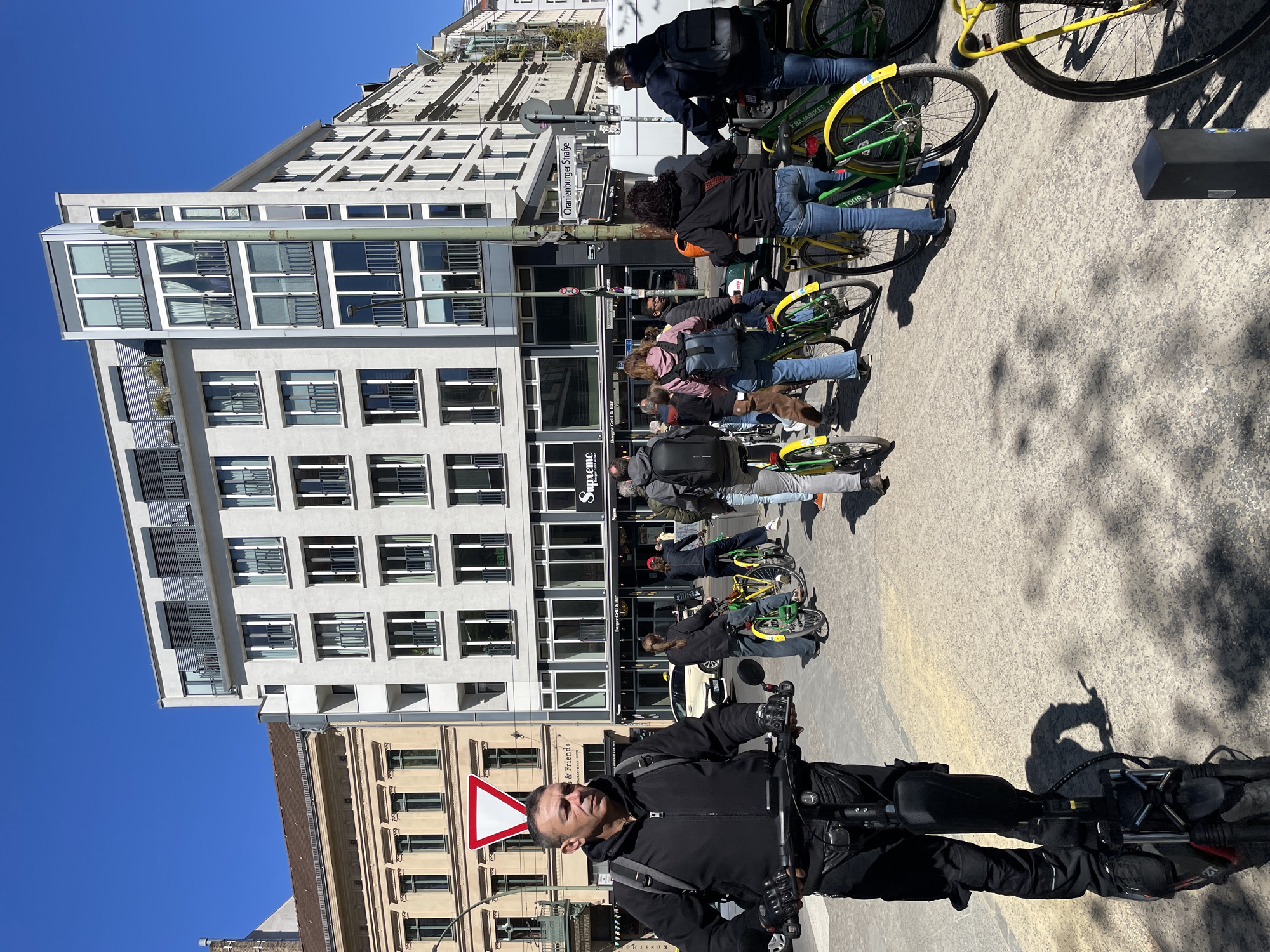 The image depicts an urban street scene on a bright, sunny day with a clear blue sky. A group of people, primarily adults, are gathered on a wide paved area, mostly standing next to bicycles. Many of the bicycles are yellow and green and branded "Bajabikes Tour," suggesting a guided bike tour or a group outing.
In the foreground, a man in a black jacket and backpack is visible, holding the handlebars of what appears to be an electric bicycle or scooter, looking towards the viewer. His shadow is cast prominently on the sunlit pavement. Behind him, other individuals are engaged in conversation or waiting by their bikes, which are lined up along the street.
The background features a mix of architecture. On the left, a modern, multi-story white building with numerous windows and balconies stands tall. Further down the street, older, more traditional buildings with decorative elements are visible. The bright sunlight creates distinct shadows on the ground, indicating it is likely mid-day.
Several pieces of text are visible, providing location cues and identifying businesses:
- A street sign clearly reads "Oranienburger Straße," confirming the location as Berlin, Germany.
- Shopfronts display names such as "Supreme," "Burger Grill & Bar," and part of another sign that says "Friends."
- The bicycles have "Bajabikes Tour" branding.
- A red and white triangular yield sign is visible in the lower-left.
The scene conveys a sense of activity and leisure, with participants appearing relaxed and ready for their bicycle journey.