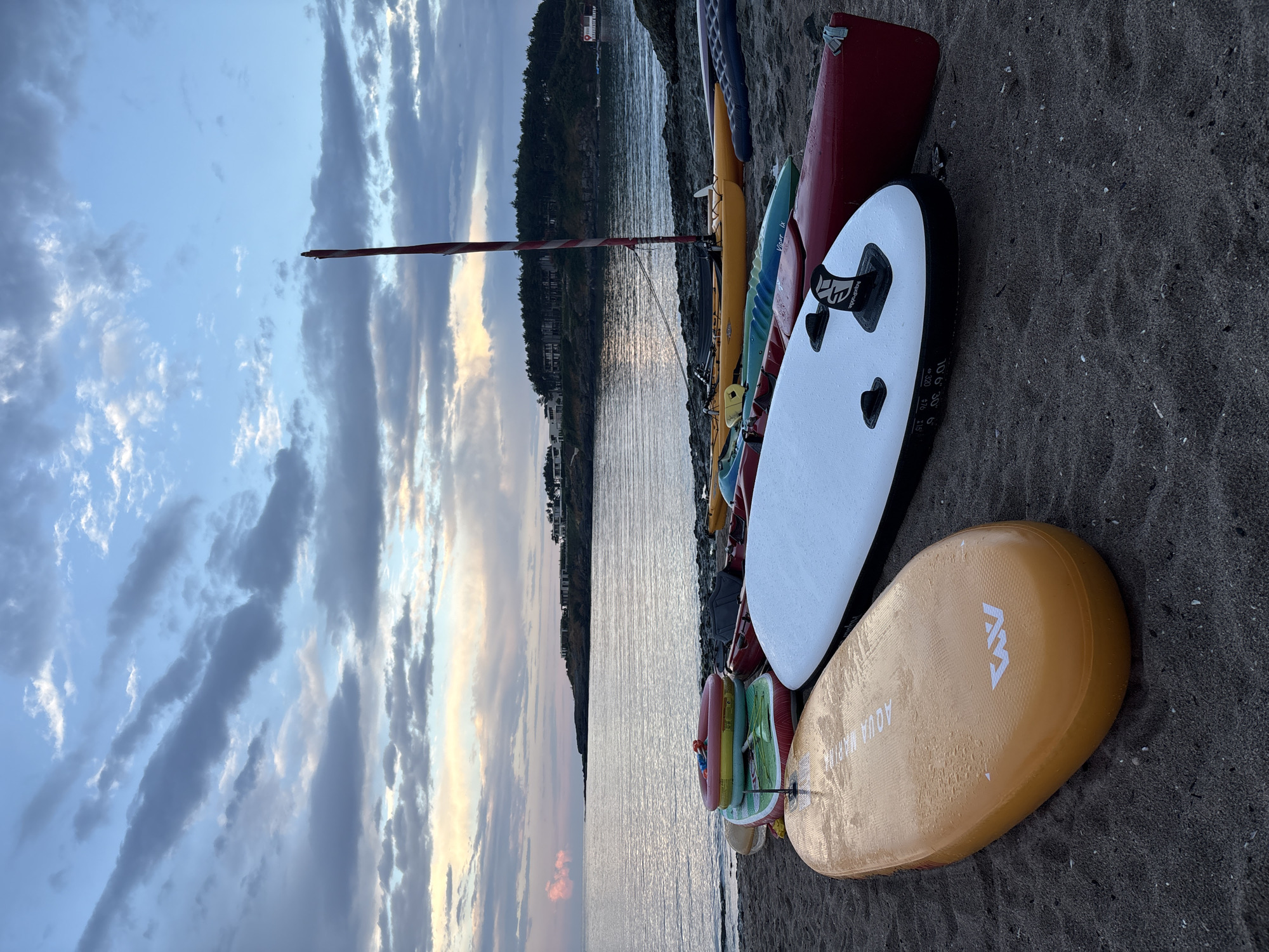 At dusk on a beach in Tsarevo, Bulgaria, various watercraft are staged on the sand. These include multiple paddleboards, kayaks, and an inflatable dinghy. A mast from a small sailboat stands prominently among the vessels. The sea is calm, reflecting the soft, diffused light of the setting sun, with a distant treeline and buildings dotting the shoreline. The sky is a blend of blues and grays, with wispy clouds suggesting the end of the day. The overall scene is tranquil, depicting water sports equipment resting after use or awaiting the next day's activities.