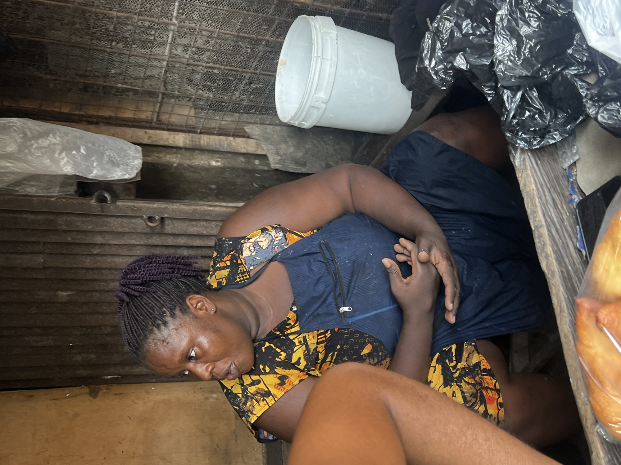 A woman sits in what appears to be a cramped, enclosed space, likely a shop or storage area in Unknown, Unknown. She wears a blue dress with a yellow and black patterned overlay. The background is cluttered, with a white bucket, black plastic bags, and wooden planks. The overall impression is one of confinement and possibly hardship.