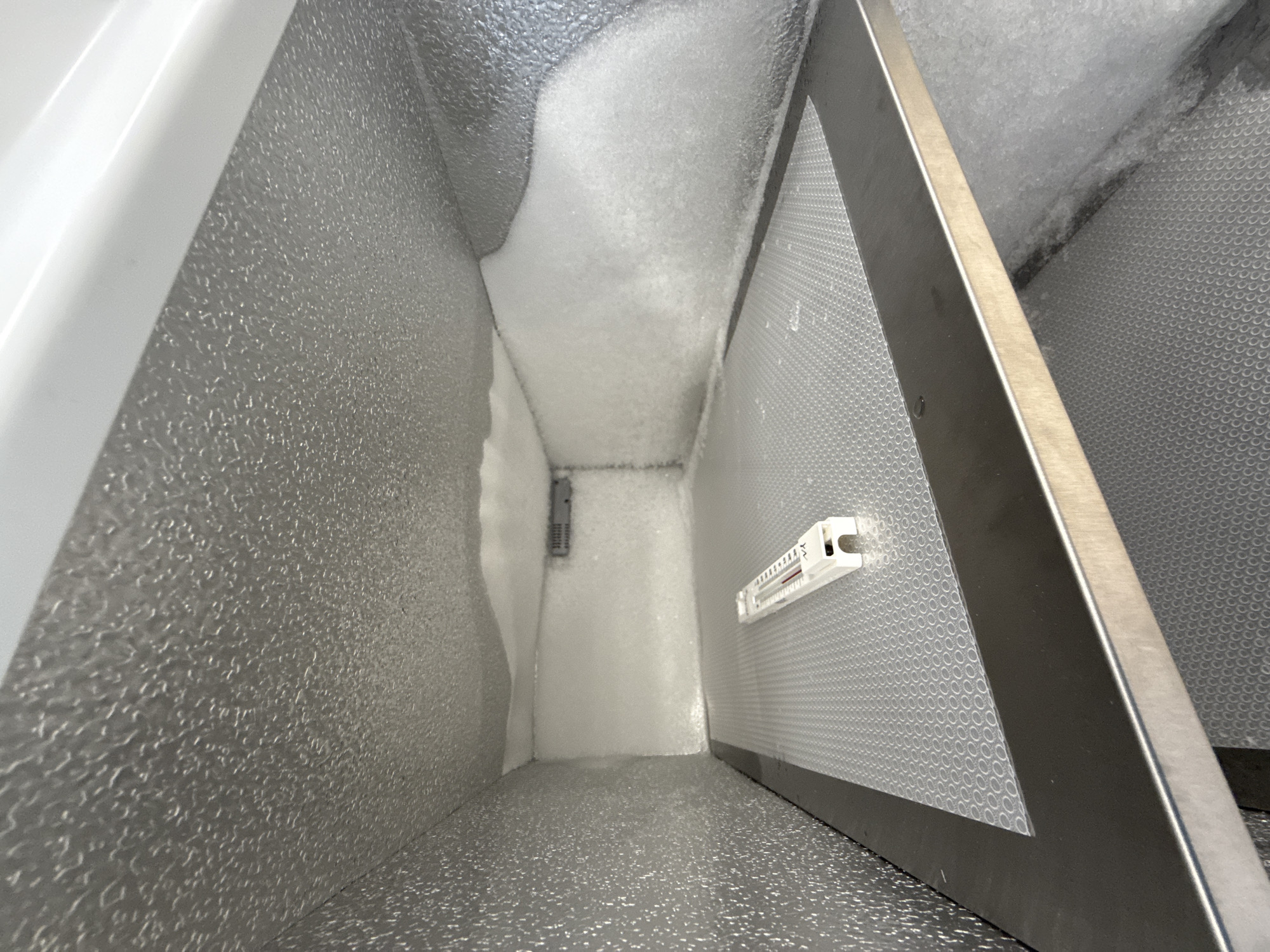 The image captures the interior of a commercial-grade freezer or cold storage unit in Moscow, Russia, viewed from a slightly elevated perspective looking down its length. The walls and floor are constructed from a light gray, textured material, possibly aluminum, exhibiting a hammered or dimpled pattern.
Significant frost and ice buildup are visible, particularly on the upper sections of the walls and along the back wall, indicating the cold operational environment of the unit. The frost appears as a thick, powdery white layer.
On the back wall, a small, dark gray electronic component, possibly a sensor or part of a temperature control system, is partially obscured by the frost. To the right, a vertical panel with a fine, perforated or honeycomb pattern is present, framed by a stainless steel edge. Attached to this panel is a white plastic device, resembling a thermometer, with a red liquid column and numerical markings (e.g., "10", "20", "30") and what appears to be a degree Celsius symbol ("C"). There are also small triangular arrow symbols ("^^") at the top of this device.
No human subjects or activity are depicted. The scene suggests a static, functional environment of a cold storage facility. The lighting is even, illuminating the interior and highlighting the textures and frost.