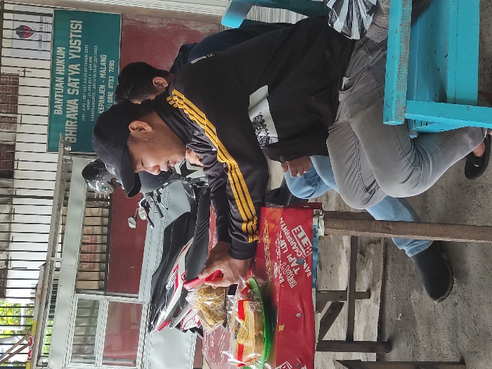 This outdoor image captures a casual street scene featuring two men and various objects under bright daylight. In the foreground, a man wearing a black long-sleeved shirt with yellow stripes, a black cap, and grey jeans stands, seemingly arranging or handling items on a small wooden stand. His hands are engaged with several clear plastic bags containing snacks and a red package, which displays fragmented text like "RERAT", "TAPI LUP", and "RETE". Behind him, a black and white motorcycle is parked.

To the right, another man is partially visible, seated on a bright blue wooden chair, wearing a light blue shirt and jeans. The ground appears to be concrete or pavement. In the background, mounted on a building wall, is a prominent green sign. The sign identifies a legal aid service: "BANTUAN HUKUM BHIRAWA SATYA YUSTISI", followed by registration numbers, "TAKESHANGPOL: 230/410/35.07.207/2023", "KEPANJEN - MALANG", and a contact number "0812 8793 7295". The scene suggests an everyday interaction, possibly involving street vending or distribution in Kepanjen, Indonesia.