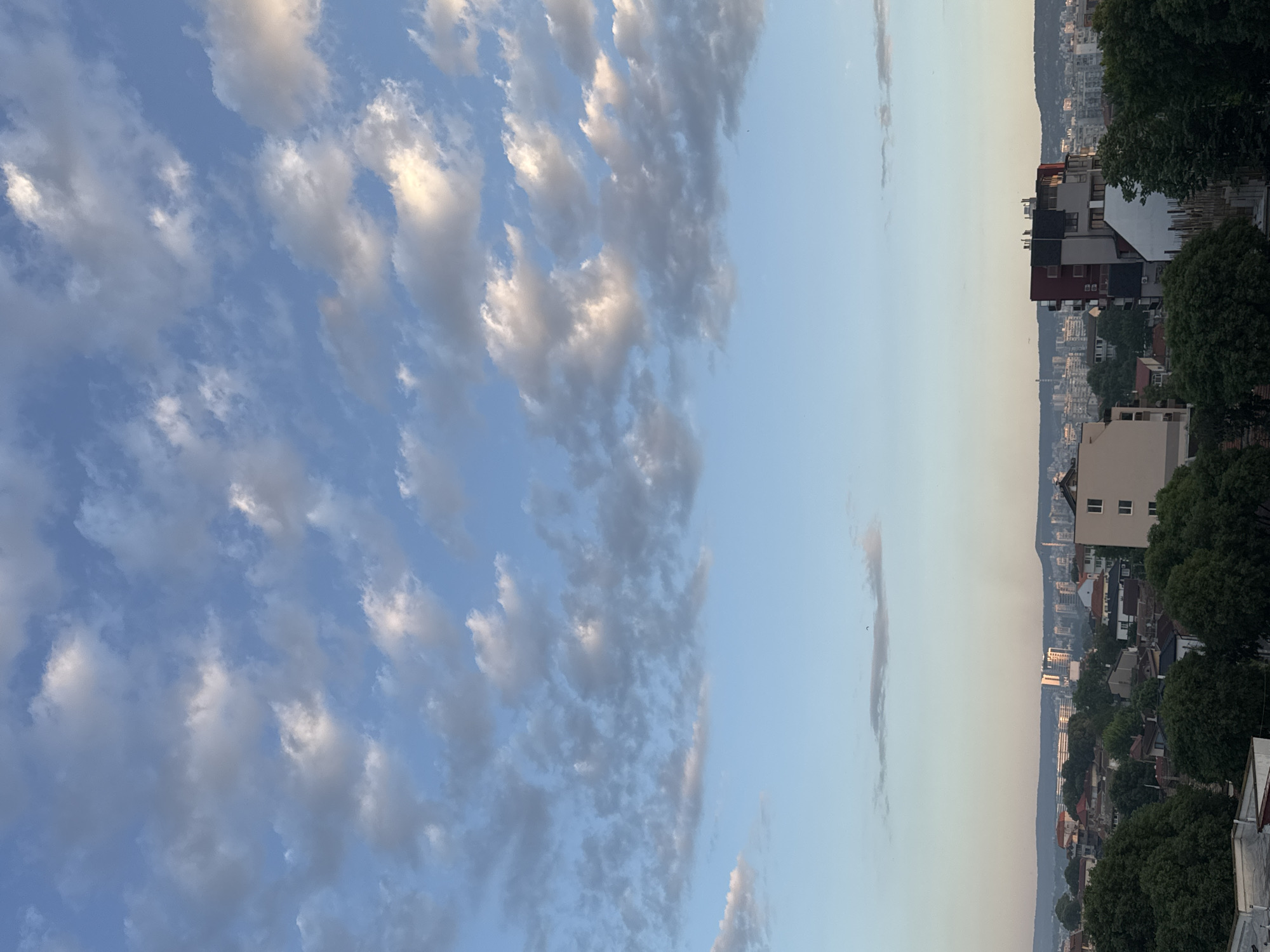 The image shows a cityscape in Varna, Bulgaria, under a sky with altocumulus clouds. The right side of the image reveals buildings of varying heights and colors, interspersed with trees. A narrow strip of clear sky separates the clouds from the urban landscape. In the background, more buildings and possibly distant hills are visible through a slight haze. The light suggests it is either early morning or late afternoon.