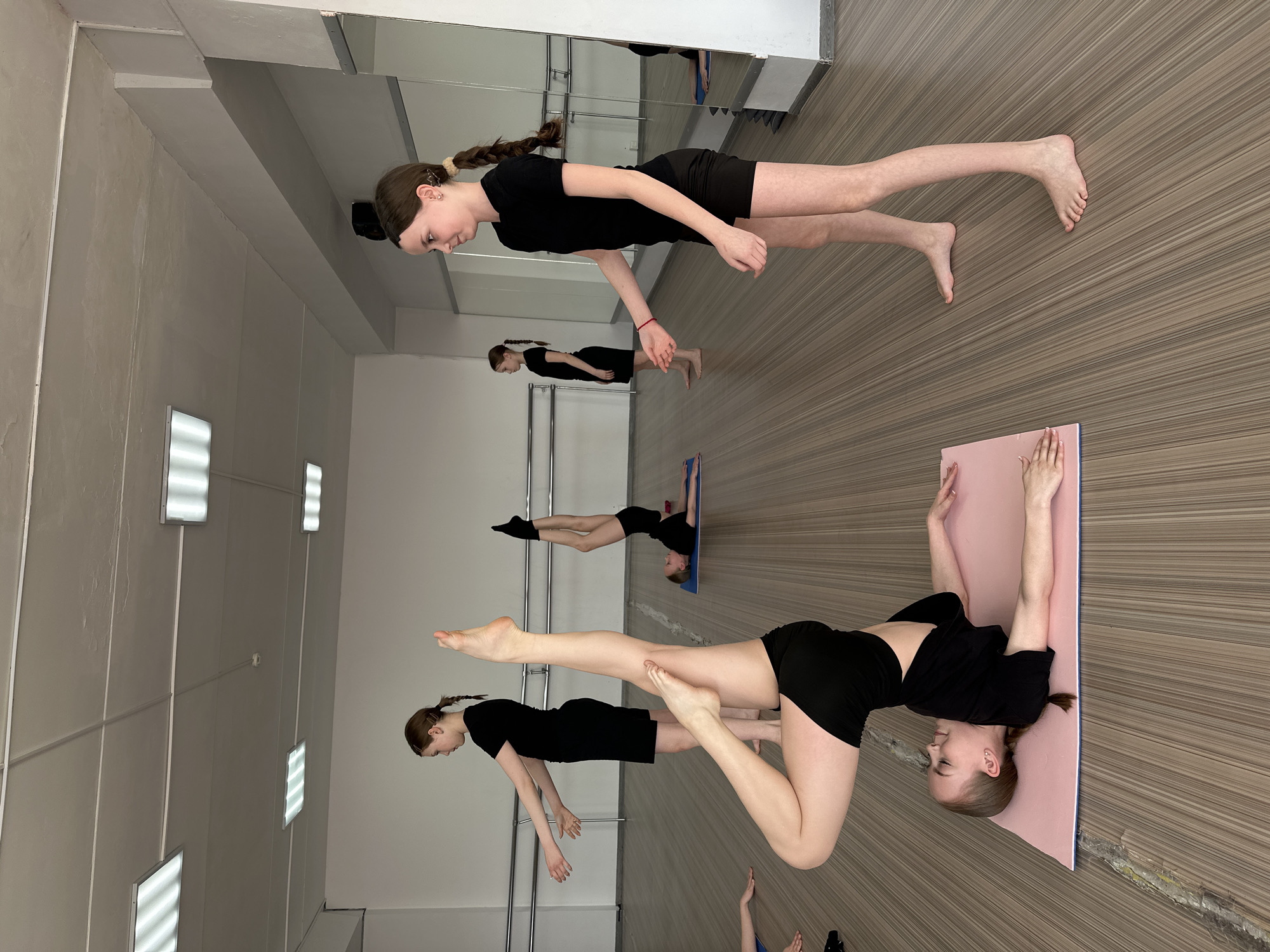 The image displays an indoor physical activity session in Votkinsk, Russia, featuring five young females. The studio is characterized by a light-colored ceiling with multiple rectangular fluorescent light fixtures, light-colored walls, and a striped brown floor. A large mirrored wall on the right side of the room contains two horizontal ballet barres and reflects parts of the interior and individuals. In the foreground, one female lies supine on a pink exercise mat, performing a modified shoulder stand with her legs extended vertically and slightly bent at the knees. To her immediate right, another female stands upright, facing left, with both arms extended forward and palms open. In the mid-ground, a third female stands near a ballet barre, also with arms extended forward. Adjacent to her, a fourth female lies supine on a blue exercise mat, executing a similar inverted leg raise. All participants are barefoot and dressed in matching black t-shirts and black shorts. The hands and lower arms of a fifth individual are partially visible in the bottom left corner of the frame.