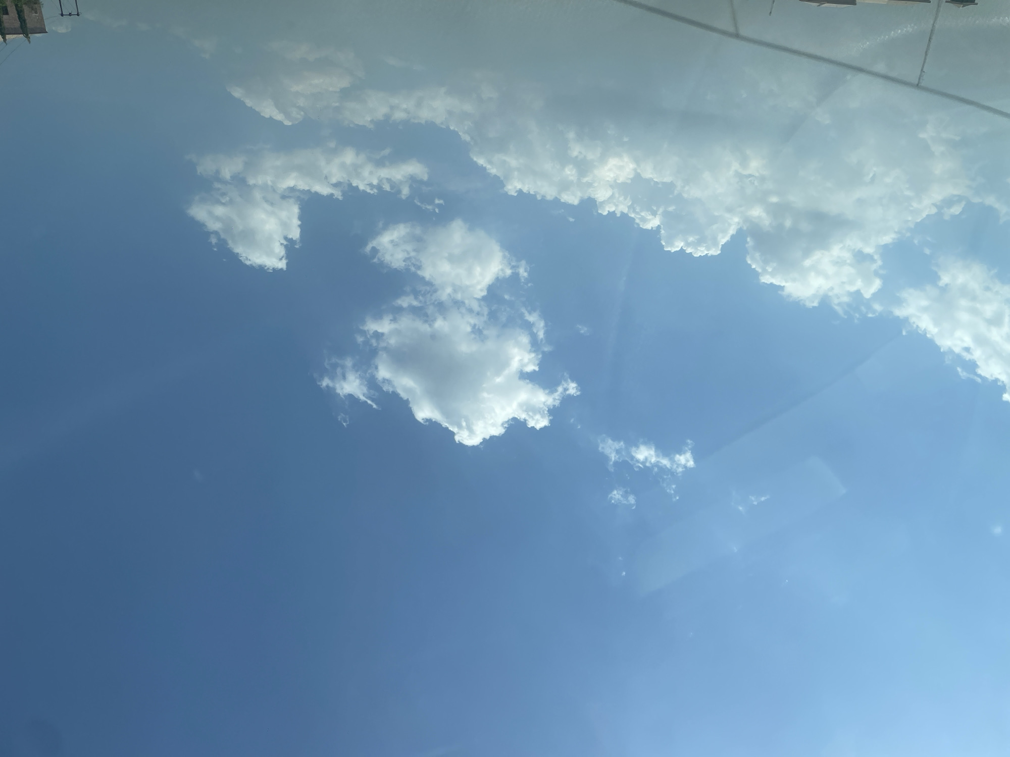 The image captures a bright, expansive blue sky with scattered white clouds, likely taken through a window, possibly from inside a vehicle or building. The primary subject is the sky, dominated by vibrant blue hues.
Numerous cumulus clouds are visible, ranging from small, wispy formations to larger, more defined, fluffy masses. A prominent band of denser white clouds stretches across the upper portion of the frame (when rotated to a landscape orientation). The clouds are brightly lit, suggesting direct sunlight.
Reflections on the glass surface are noticeable, particularly in the lower right and along the top edge, indicating the camera's position. These reflections create faint, indistinct shapes and lines. Glimpses of what appear to be the tops of distant structures or buildings, along with power lines or poles, are visible in the upper left corner through the glass or as reflections. In the top right, faint linear patterns suggest either a road or architectural details.
The scene depicts a calm, clear daytime environment, indicative of fair weather. The bright illumination of the clouds and sky points to a time of day when the sun is high, likely late morning or afternoon. No specific activity or event is taking place, other than the observation of the sky. There is no visible text in the image. Given the provided context, the image captures a sky view from Crescent, Nigeria, though no specific visual cues within the image confirm this location.