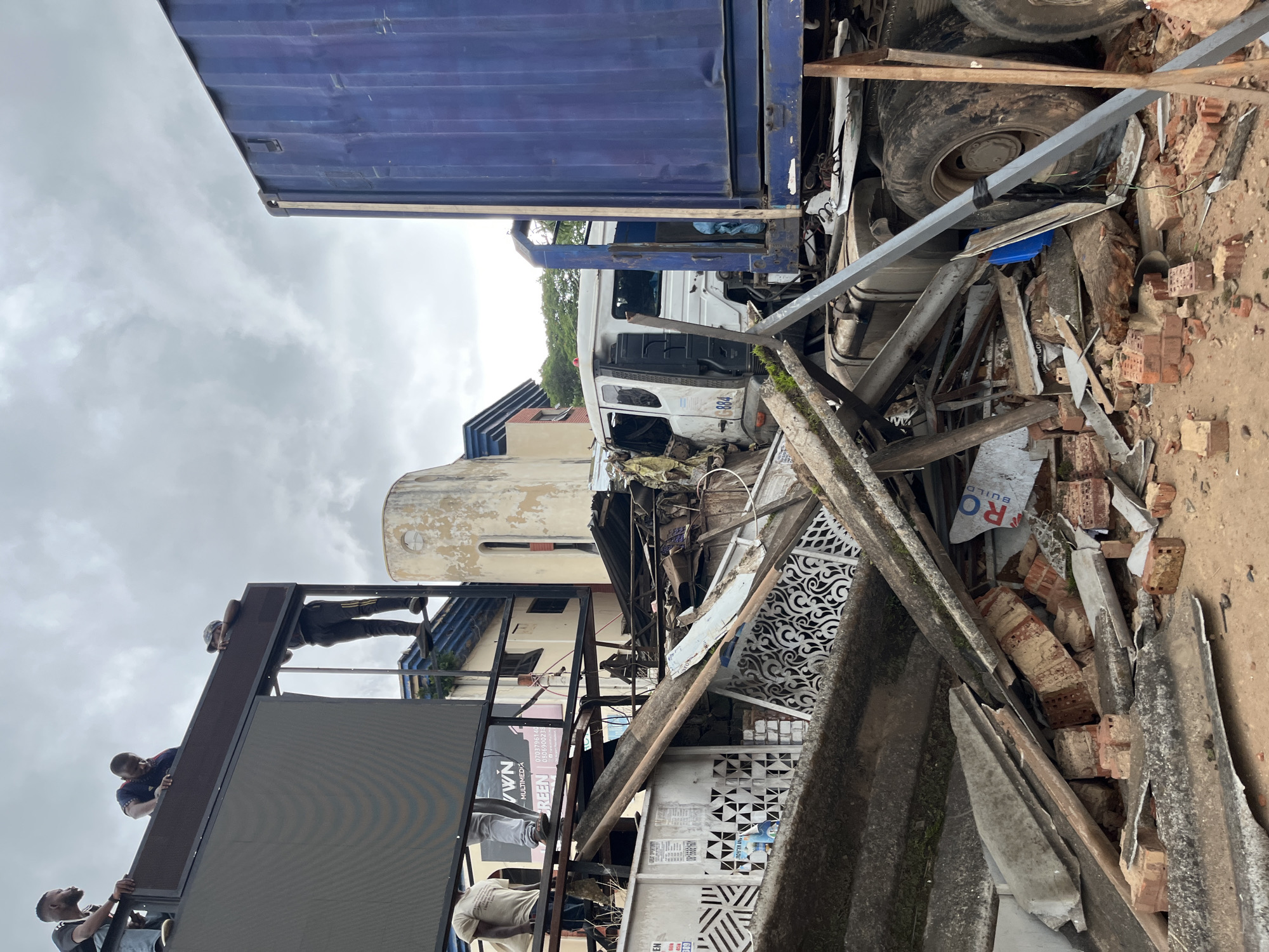 In Barakin Lerawi, Nigeria, an image depicts the aftermath of a significant structural collapse. A large, blue shipping container truck is positioned on the right, its trailer partially obscured by debris. To the left of the truck, a white truck cab shows visible damage, with its front end crushed and its windows shattered. The scene is littered with rubble, including bricks, corrugated metal sheets, wooden beams, and concrete fragments. In the background, a large, dark digital billboard, possibly a jumbotron, is elevated on a metal frame. Several individuals are visible near the billboard, some standing on its platform and others on adjacent structures. One person, wearing a dark shirt and a cap, is on the billboard platform, appearing to be working. Another person in a blue and red patterned shirt is also on the platform, leaning over. Further to the left, two individuals are on a higher platform, one in a dark shirt and the other in a lighter shirt. The overall environment suggests a chaotic and damaged urban setting under an overcast sky.