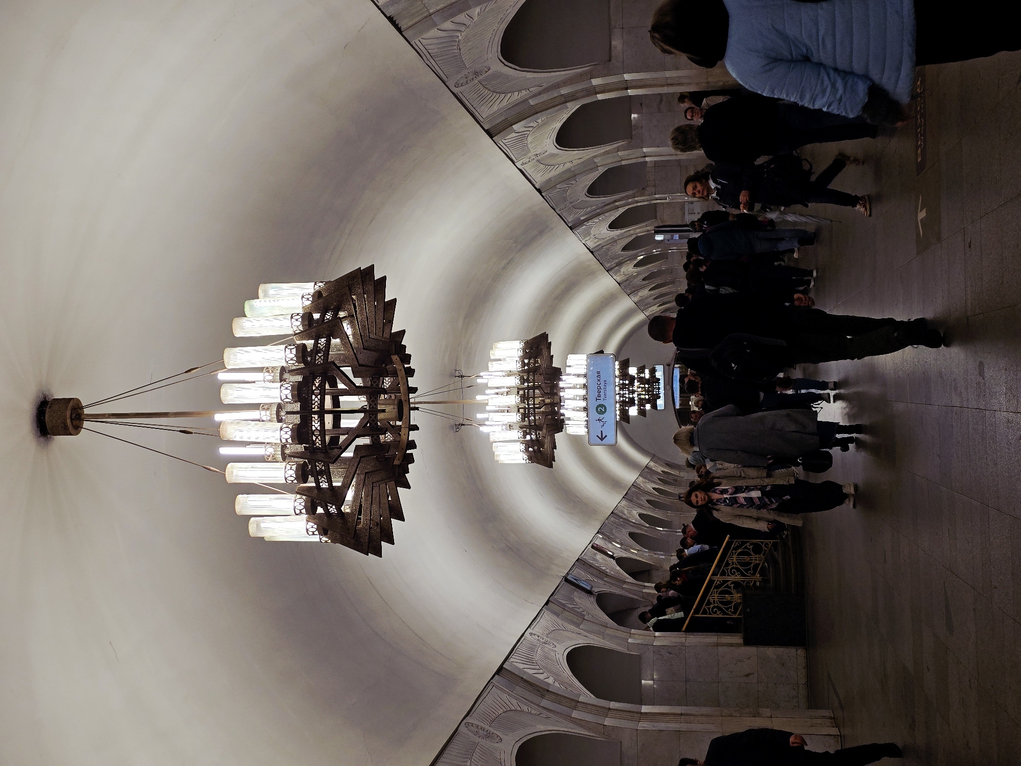 The image shows a view inside a Moscow metro station, Russia. In the foreground hangs a large, ornate chandelier with multiple light sources. The station has a curved ceiling with arched details along the side walls. People are walking and standing on the platform, and there is a sign indicating the direction to "Тверская" (Tverskaya) station.