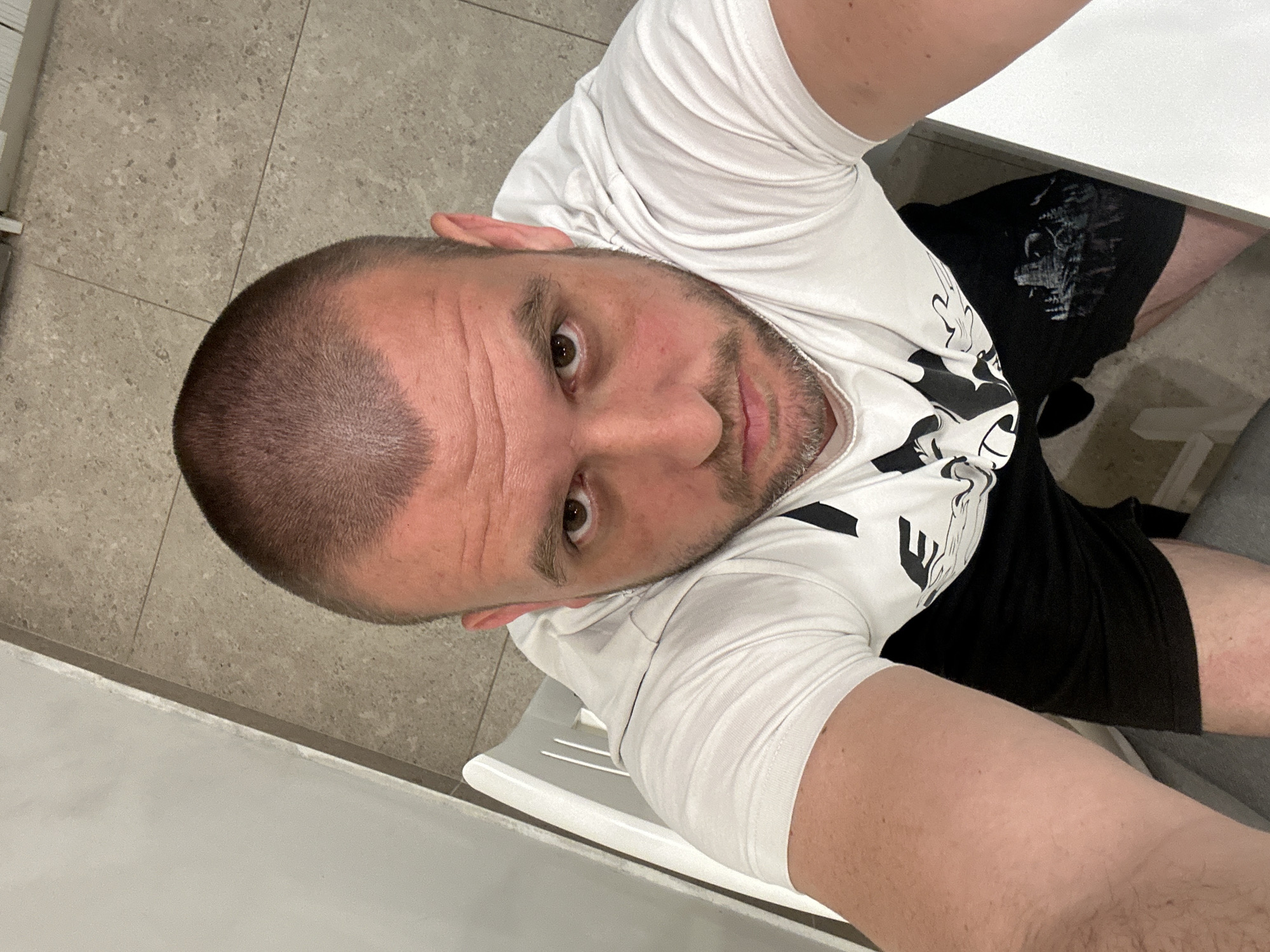 A man is sitting indoors, likely in an apartment or office. He is fair-skinned with a shaved head and a trimmed beard. He is wearing a white t-shirt with a design and black shorts. He is seated on a white plastic chair and the floor is tiled with a grey stone. Part of a white table is visible to his right. The location of the image is unknown, so it's not possible to determine if this image was taken in Sofia, Bulgaria.