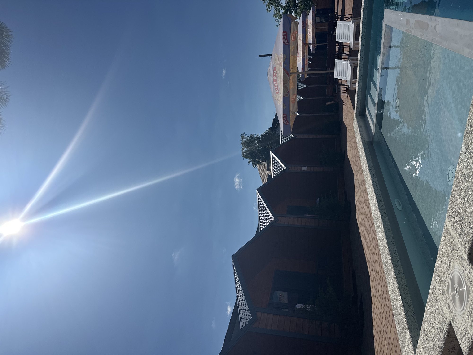 This is a sun-drenched outdoor scene, likely a resort or hotel complex, with small wooden cabins lined up on the left, each with a dark door and what appears to be a number plate visible on one. In the foreground, a section of a swimming pool is visible, with steps leading into clear blue water. Two white lounge chairs are positioned beside the pool. Several large, open umbrellas are set up further back, providing shade over tables and chairs, with branding that appears to read "PULPY". The sky is a clear, bright blue with a prominent sun flare creating streaks of light across the image. The ground is paved with red brickwork. The overall impression is one of a warm, sunny day at a recreational facility.