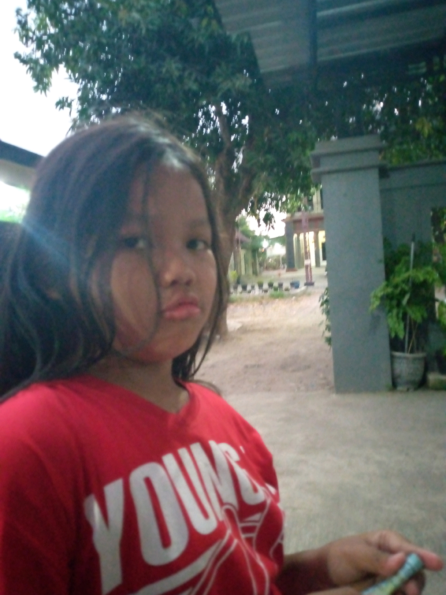 A young girl with long, dark hair is pictured from the chest up, wearing a red t-shirt with white lettering that reads "YOUNG." She is looking directly at the camera with a neutral expression. Her hair is slightly messy, falling around her face and shoulders. The background is slightly blurred, suggesting an outdoor setting. Visible elements include trees with green foliage, a paved or concrete ground surface, and a gray concrete pillar on the right side of the frame. The overall lighting suggests it is daytime, possibly overcast. The scene appears to be in the city of Sumberwono, Indonesia, based on the available information.
