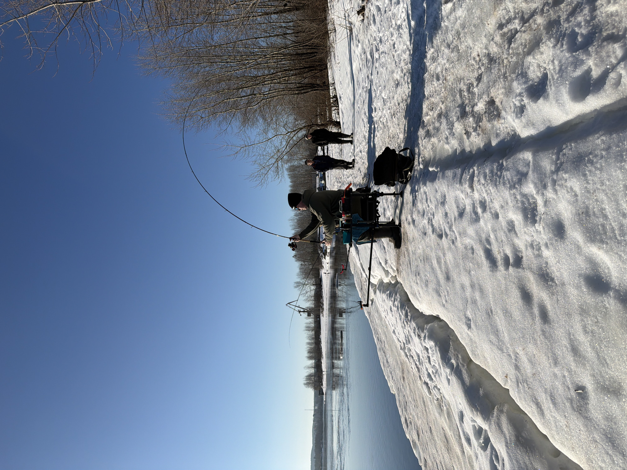 The image depicts a man fishing in winter. He stands on a snow-covered bank next to a body of water, likely a river or lake. He is wearing cold-weather gear, including a hat and waders, and holding a fishing rod. The rod is bent, suggesting a fish is on the line.
In the background, there are bare trees lining the opposite bank. The sky is clear and blue. There are a few other people standing behind the fisherman.
A crane is seen on the water, presumably connected to construction.
Based on the information provided, the scene is located in the city of Noviy, Russia.