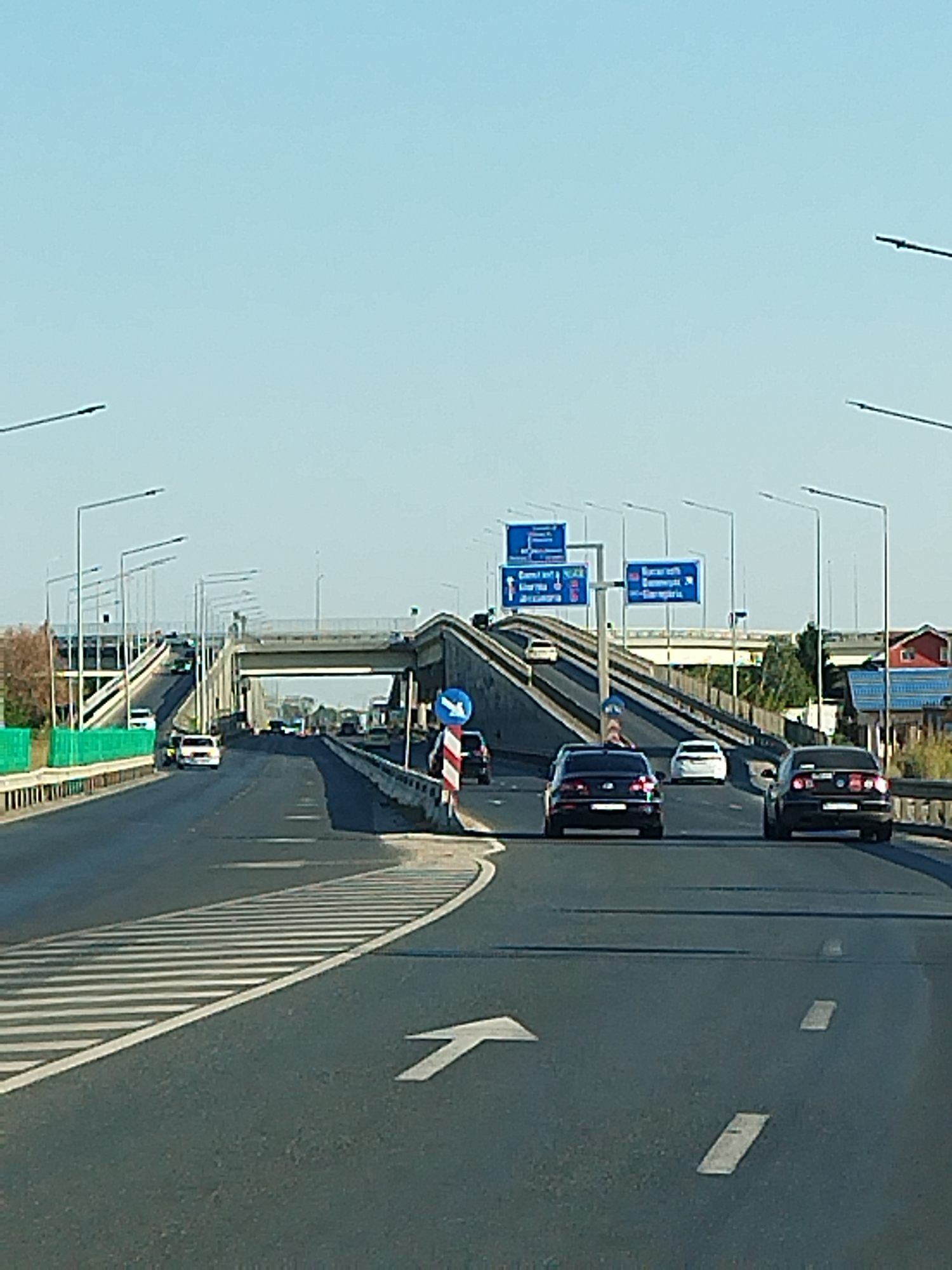 The image depicts a multi-lane highway on a clear day, likely in or near Olteni, Romania, indicated by road signs. Several vehicles are visible, including black sedans and white cars, traveling in both directions. The road features clear lane markings, including white arrows indicating traffic flow. Overhead bridges and streetlights are prominent architectural elements. On the right side of the road, a blue directional sign with white text points towards "Constanta" and "Alexandria," and another sign with "Bucuresti" and "Giurgiu." A striped pole with a blue circular sign displaying a white arrow is also present. The environment appears to be a well-maintained highway infrastructure.