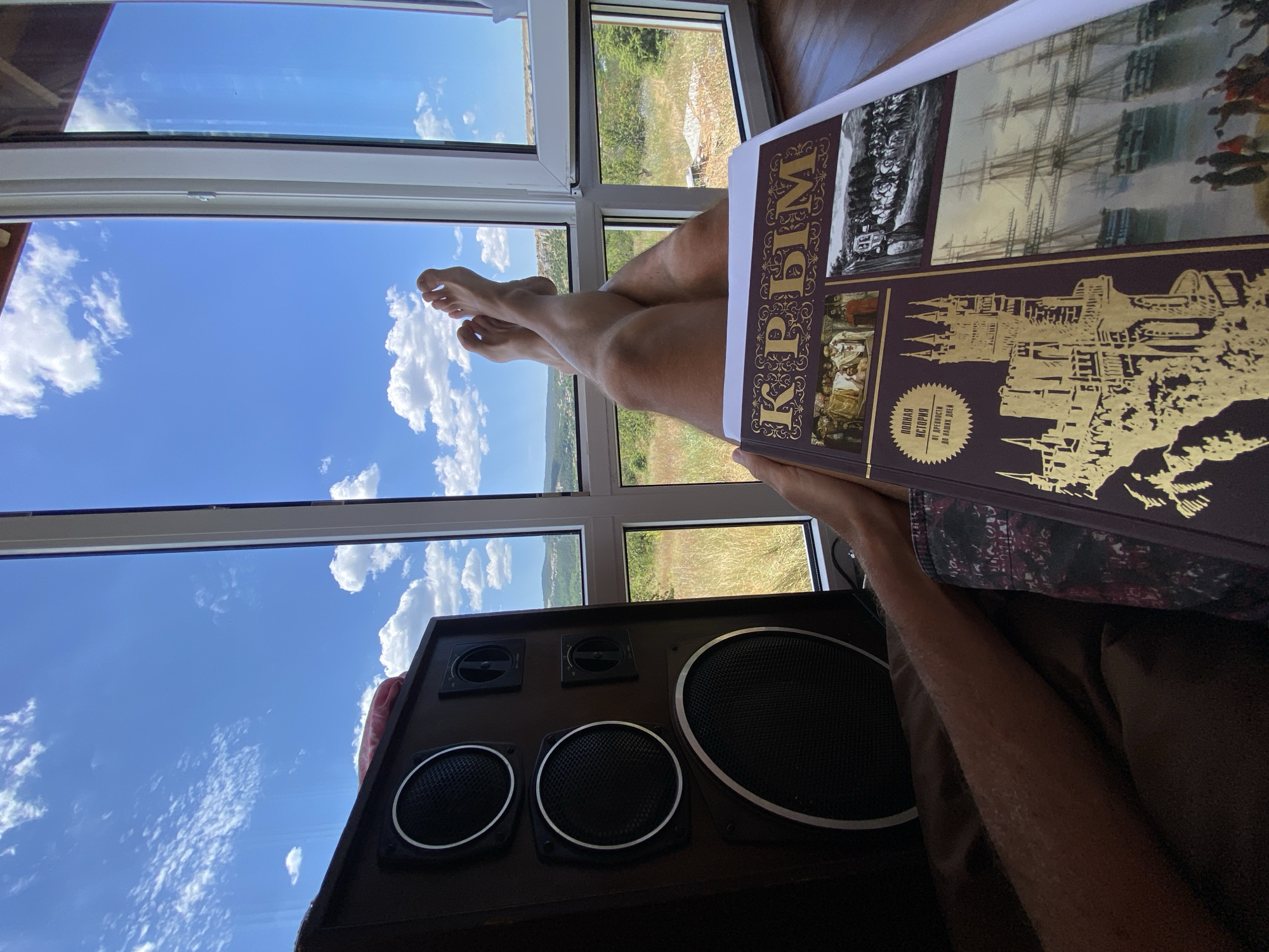 A person is lying on their back, with their legs crossed and feet propped up near a window. They are holding an open book with a dark cover that has gold lettering and illustrations. The book appears to be about history, with images of historical figures, a castle, and ships.
The person is wearing patterned shorts. Their legs are tanned and slightly hairy. A large, dark brown speaker is positioned to the left of the frame, partially obscuring the person's arm.
The scene takes place indoors, near a large window that offers a view of a rural landscape. The sky is blue with scattered white clouds. The landscape outside consists of dry grass, trees, and hills in the distance, with some buildings visible. The sunlight indicates it is daytime. The location is described as Krasnomakskoye rural settlement, Ukraine.