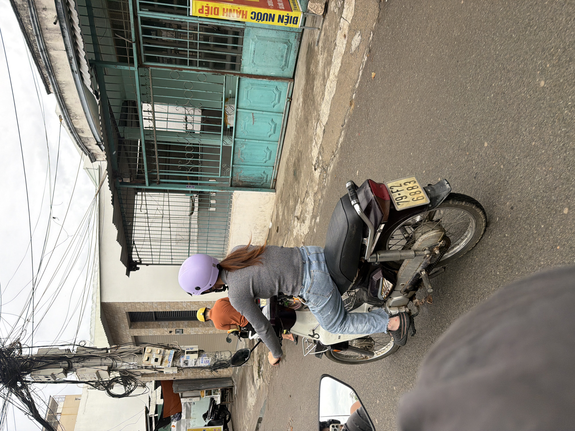 An overcast day in Nha Trang, Vietnam, is captured from the perspective of a rider on a motorcycle, looking forward. The primary subject is a female rider wearing a light purple helmet and a gray long-sleeved shirt with blue jeans, seated on a dark-colored scooter. She is in motion, with her left hand on the handlebar. Behind her, another person wearing an orange shirt and a yellow helmet is also on a scooter, traveling in the same direction. The street is paved, with a visible curb and sidewalk. Buildings line the street, featuring a mix of architectural styles. One prominent building has a turquoise metal gate and a weathered, light-colored facade. Another building appears to be under construction or in a state of disrepair, with exposed wires from a utility pole dominating the background. A yellow sign with Vietnamese text is visible on the right side of the image. The overall scene depicts a typical urban streetscape in Vietnam, characterized by the prevalence of motorcycles and a somewhat chaotic arrangement of utilities.