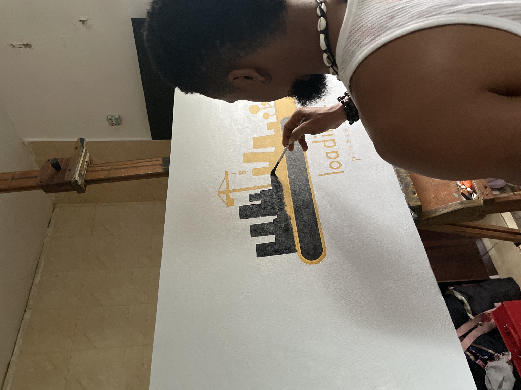 A man is painting a white canvas with a design consisting of a gold crane, gold buildings, and a gold outline of the phrase "Loading Please", using a paintbrush with black paint to fill in the buildings. He is wearing a white tank top and a black and white beaded necklace. The canvas is propped on a wooden easel. The scene appears to be indoors. The location cannot be confirmed to be in Ado, Nigeria.