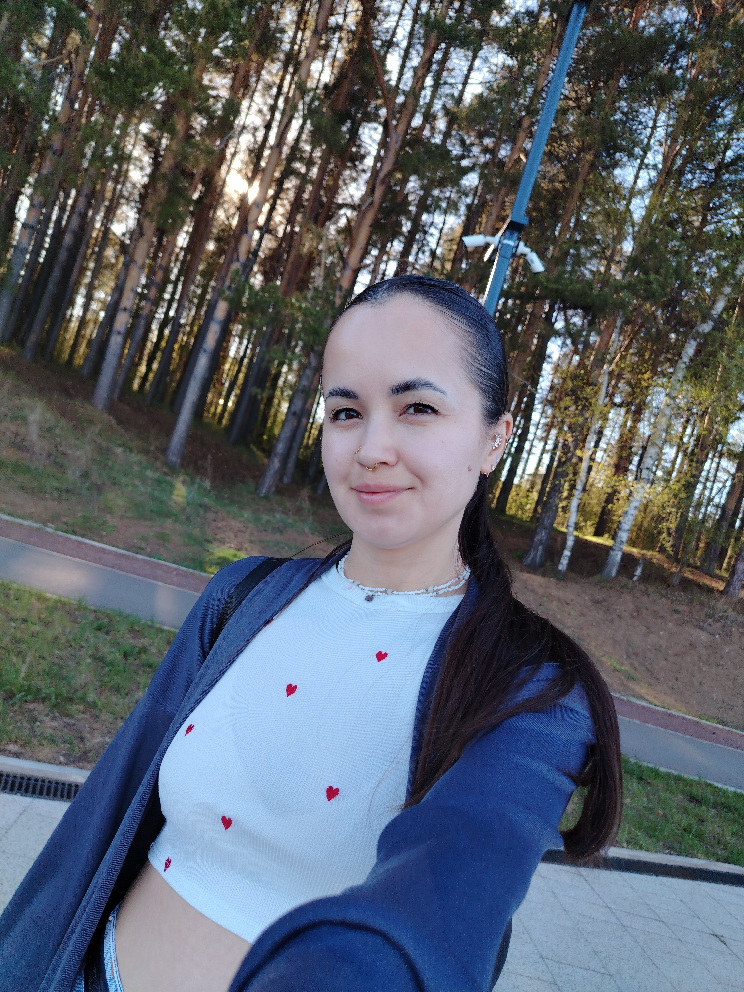 A young woman is taking a selfie outdoors in Chaikovsky, Russia. She has dark hair pulled back into a ponytail, and wears a white crop top with red hearts, a dark blue jacket, and a layered necklace. She has a nose ring and earrings. The background features a forested area with tall trees and a paved area. A lamppost is visible behind her to the right.