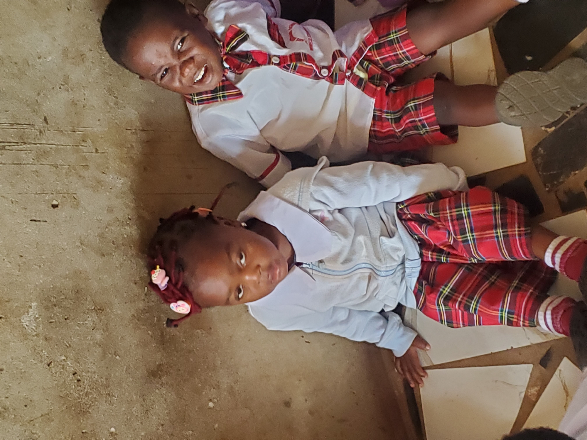 Two children are lying on a tile floor. The child on the top is wearing a white shirt with a red plaid trim and a matching bow tie and shorts. The child on the bottom is wearing a light blue jacket, a white shirt, and red plaid shorts. Both children are dark-skinned. The location appears to be a building in Ukwu-Abwa, Nigeria, although there is no further evidence to confirm that location.