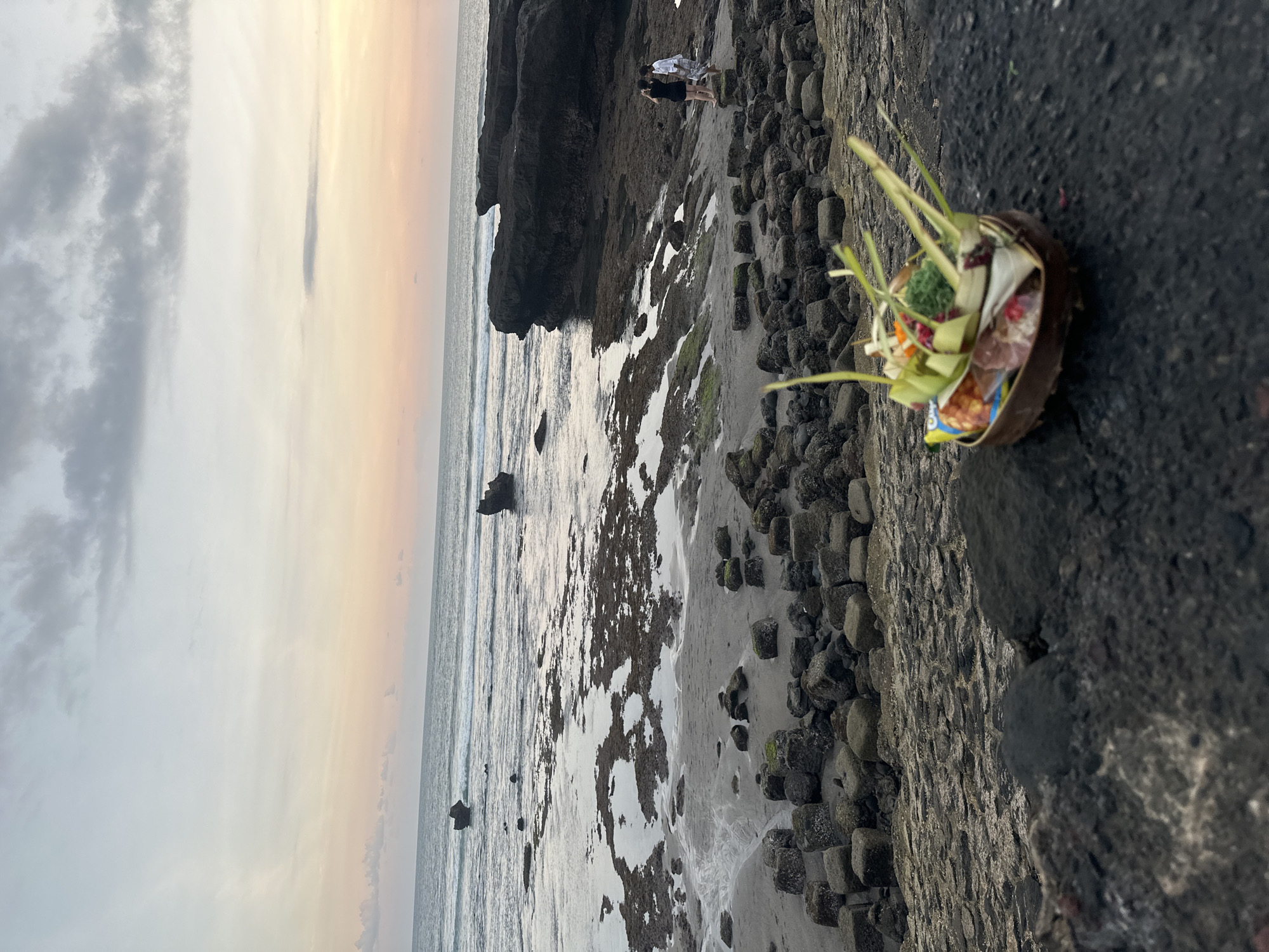 This image depicts a serene coastal scene at what appears to be either sunrise or sunset, likely in North Kuta, Indonesia, given the context.
In the immediate foreground, resting on dark, rocky ground, is a traditional Balinese offering basket, or Canang Sari. It is woven from palm leaves and filled with an array of items, including green leaves, colorful flowers, and small food offerings, signifying a cultural or religious ritual.
The mid-ground reveals a rugged coastline composed of dark, irregularly shaped rocks, some appearing like squared blocks, interspersed with patches of sand and shallow water. Algae or moss adds hints of green to some rock surfaces. Further back, two small figures of people are visible standing on the rocky terrain, near a large, dark rock formation that extends into the water. They appear to be observing the surroundings.
Beyond the immediate shore, the ocean stretches out under a vast sky. Gentle waves can be seen rolling onto the beach. The horizon is bathed in soft, warm hues of orange, pink, and yellow, blending upwards into lighter grey and white, with scattered darker clouds across the sky. The lighting is diffused, creating a peaceful and contemplative atmosphere. No text is visible in the image.