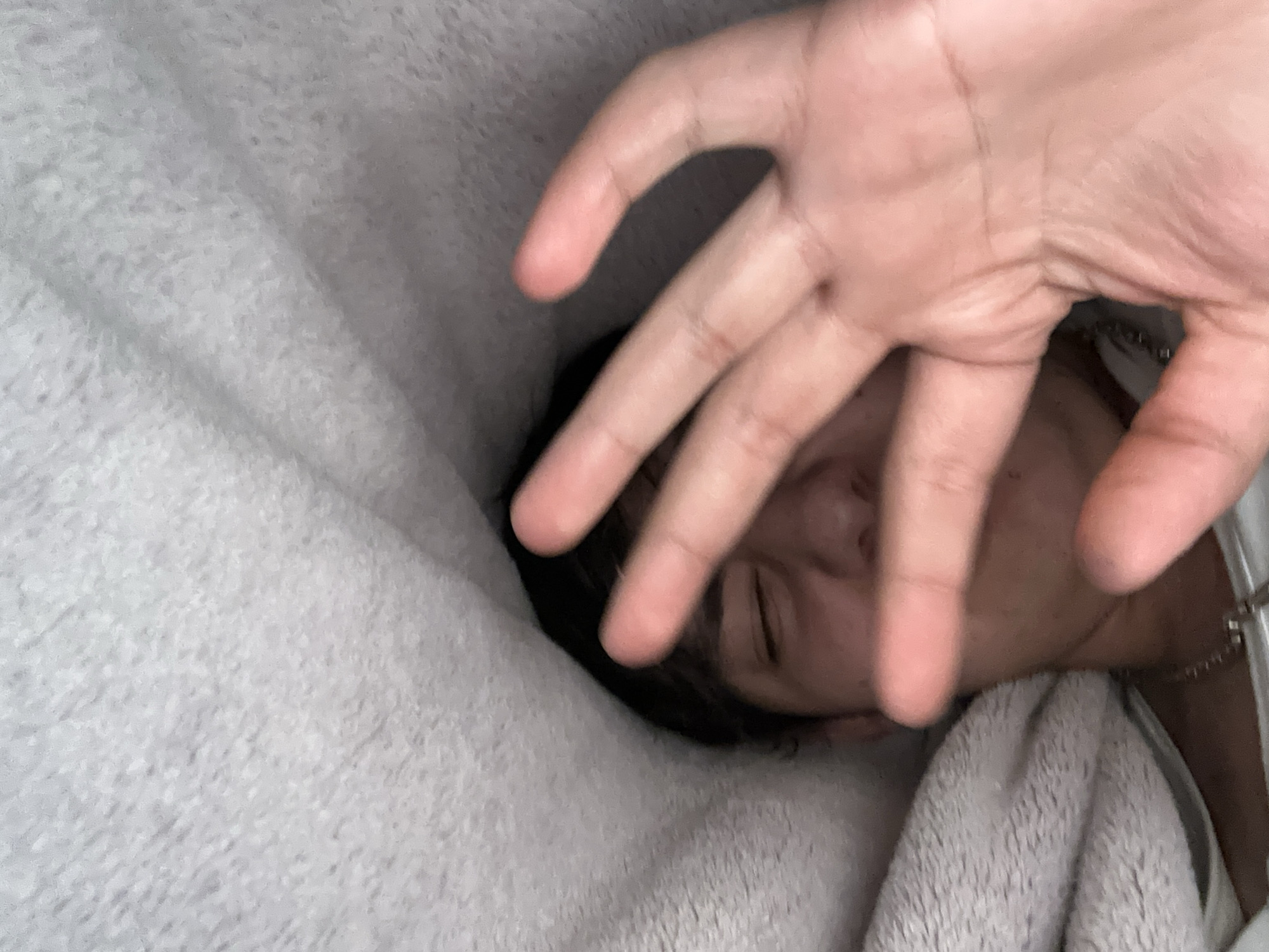 A person is lying in bed covered with a gray blanket, with their face partially obscured by a hand held up towards the camera. A silver chain is visible around the person's neck. The location is unknown.