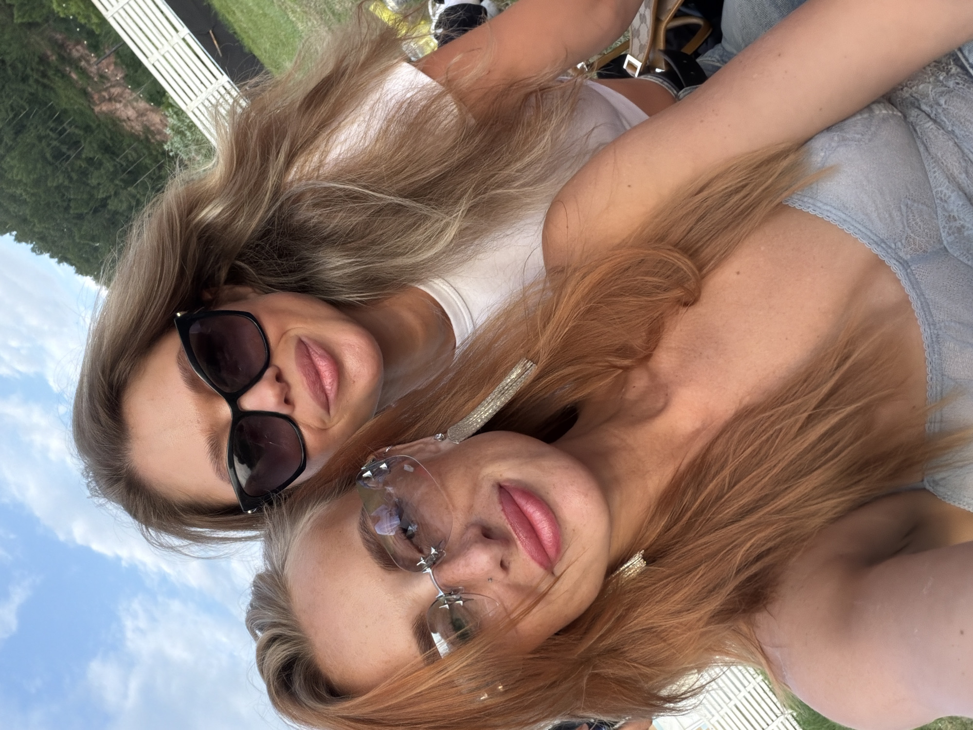 Two young women are taking a selfie outdoors in Дрокино, Russia. They are leaning in towards the camera, with their faces close together. The woman on the left has blonde hair and is wearing black sunglasses and a white top. The woman on the right has reddish-brown hair and is wearing a light blue top and clear-lensed glasses with star-shaped accents. Both women are smiling. They appear to be in a park or outdoor recreational area, with a white wooden fence and trees visible in the background. The sky is blue with some clouds, suggesting it is daytime. The overall mood is casual and joyful, indicative of a friendly outing on a pleasant day. There is no visible text in the image.