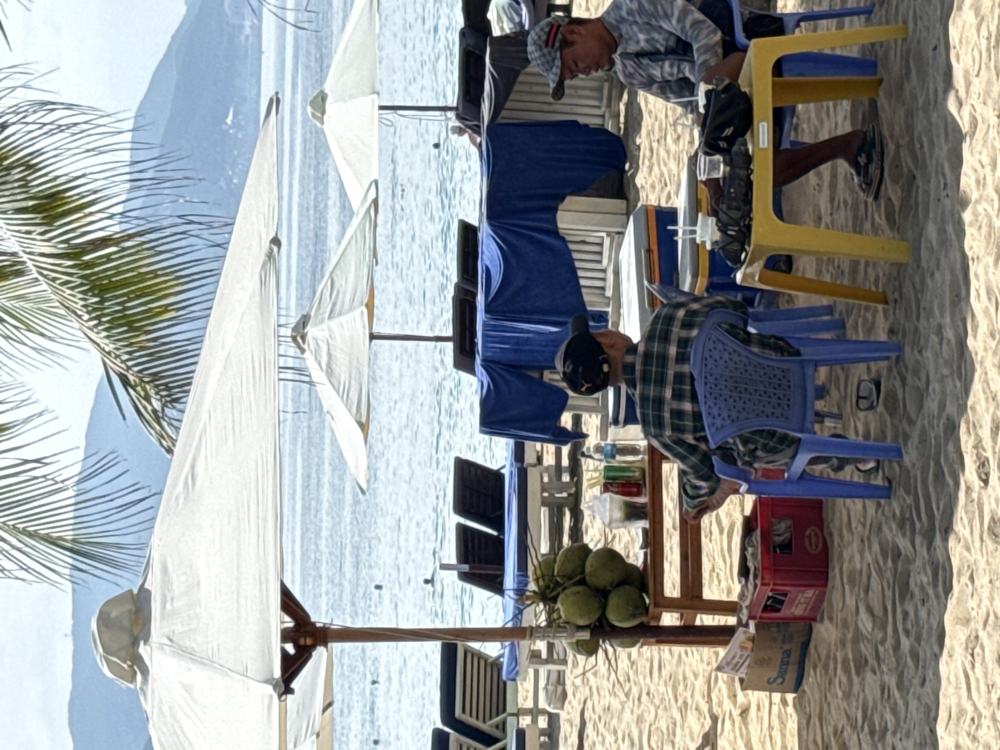 The image depicts a beach scene in Nha Trang, Vietnam, with a focus on two individuals seated at a small yellow table on the sand. In the background, the ocean stretches towards a distant mountainous coastline. Large white beach umbrellas provide shade. In the foreground, a wooden stand holds a cluster of coconuts and various beverages, including what appear to be canned drinks and bottled water, alongside cups and straws. One person, wearing a plaid shirt and a baseball cap, is seated on a blue plastic chair, facing away from the camera. The other person, wearing a patterned shirt and a checkered cap, is seated at the yellow table, looking down. The table holds several cups with straws, a black bag, and what might be binoculars. Beach chairs and a blue tarp are visible behind them, suggesting a service area for beachgoers. The sand is rippled, indicating the natural texture of the beach. The overall atmosphere suggests a relaxed, casual setting, likely a beachside vendor operation.