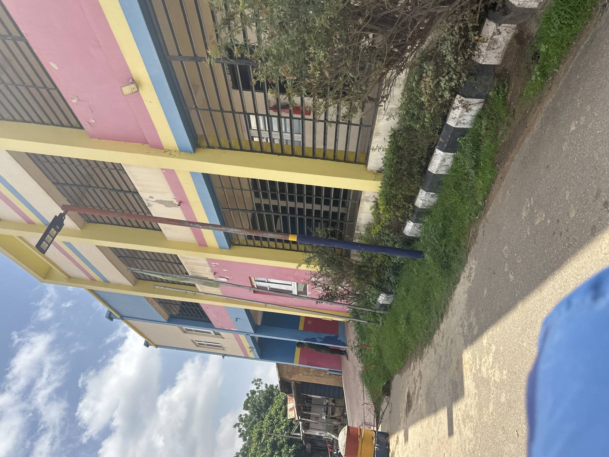 This media file is a slightly tilted, eye-level outdoor shot of a colorful building in Jos, Nigeria. The building is multi-story with bright yellow, pink, and blue sections, accented with thin blue and pink stripes. Metal grates cover many of the windows, suggesting security measures. To the right of the building, a road runs parallel to a green, grassy area bordered by a curb with alternating black and white stripes. Trees and bushes are visible, some overhanging the building and the grassy area. The sky is partly cloudy with fluffy white clouds against a blue background.
There is at least one person visible, a figure in a dark green uniform standing near the building's entrance on the ground floor, looking towards the right. The lighting suggests it is daytime, likely morning or afternoon, with shadows cast across the road. The scene appears to be a public or commercial area, possibly a school or office building, given the windows and the overall structure. The environment suggests a warm climate. No visible text is present in the image.