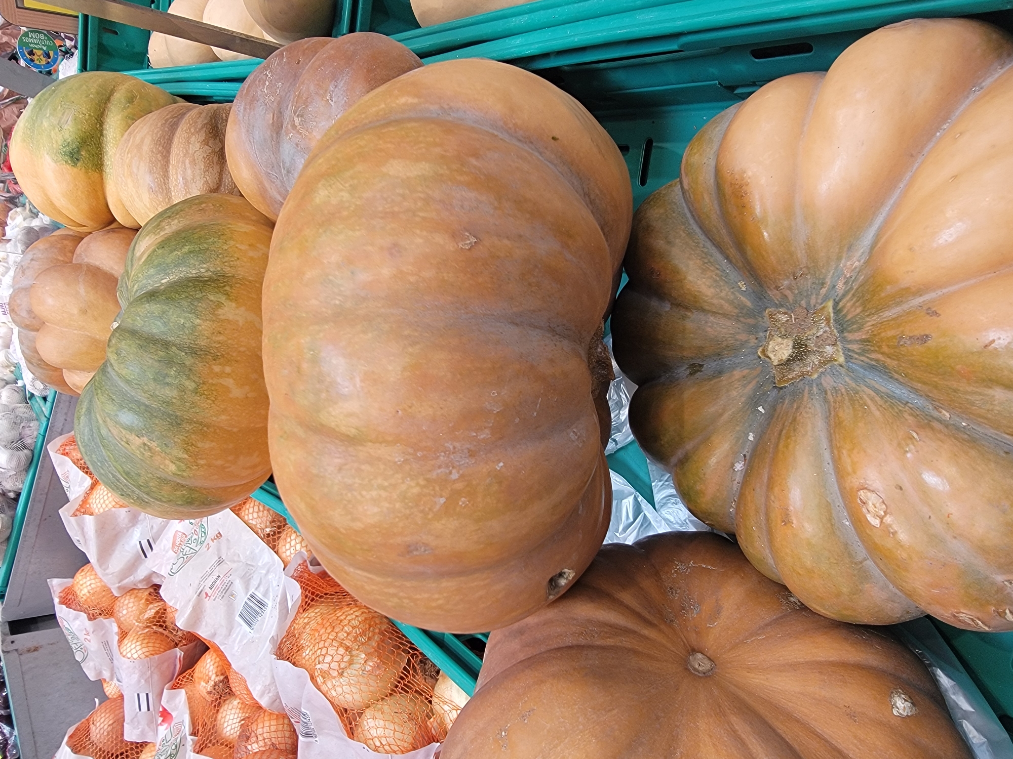A close-up, top-down view of a display of pumpkins and onions at a market in Faro, Portugal. The foreground is dominated by several large pumpkins, some with green patches and others a uniform orange-brown. One pumpkin in the center is particularly large and pale orange. Behind the pumpkins, mesh bags of onions are visible, with white packaging. The overall impression is one of fresh produce ready for sale, typical of an outdoor or indoor market setting.
