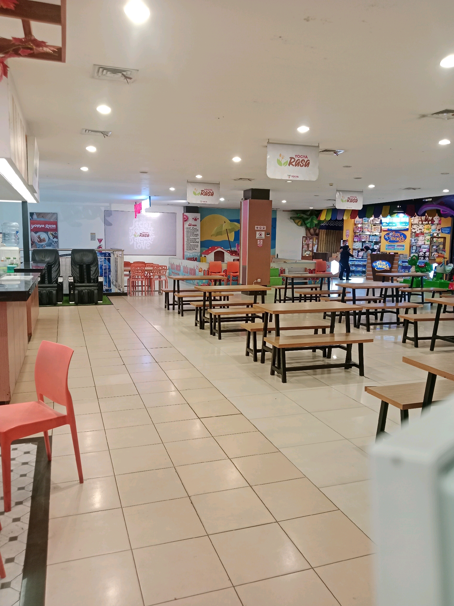 The image depicts an indoor seating area, likely a food court or a common space within a shopping mall or entertainment venue. The floor is tiled with large, light-colored square tiles. In the foreground, a bright orange plastic chair is partially visible.
The main area of the image features numerous wooden tables with black metal legs and benches. These tables are arranged in rows, suggesting a communal dining or waiting area. Some tables are empty, while others are occupied by unseen individuals.
In the background, there's a counter or service area with a black countertop. Two large, black massage chairs are placed behind this counter. Beyond the seating area, a brightly colored wall mural or display is visible, featuring cartoonish imagery and possibly advertisements. Several hanging signs are present throughout the space, some displaying text and logos, including "YOGYA RASA". A ceiling with numerous lights and air conditioning vents is also visible. The overall impression is of a clean, well-lit public space designed for customers. The location is identified as Ciranjang, Indonesia.