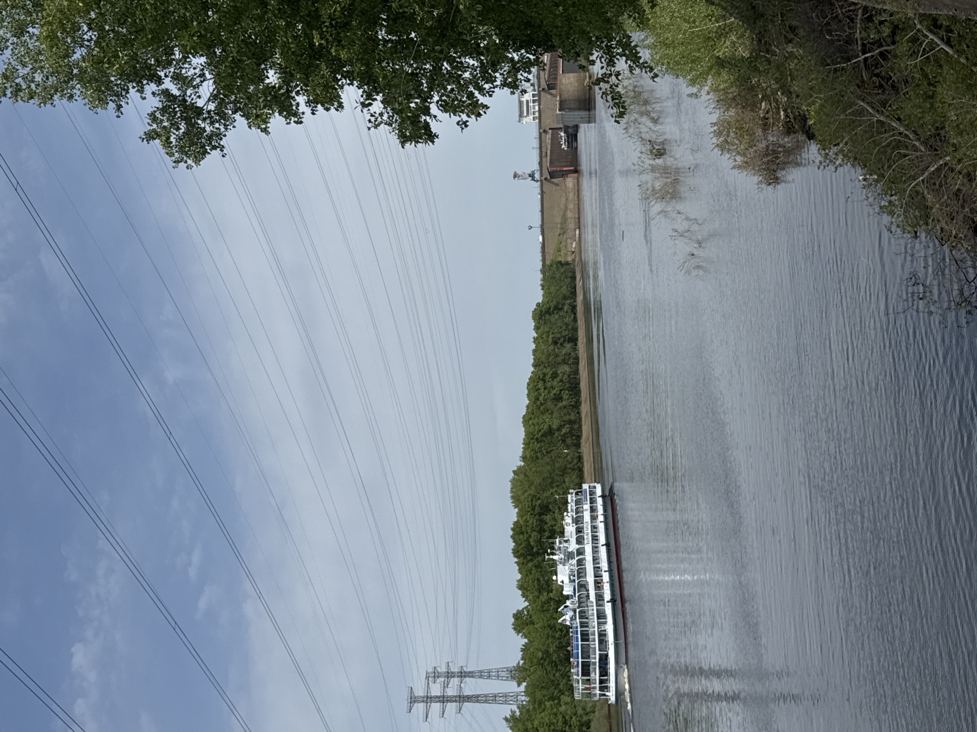 The image shows a river in Chaikovsky, Russia, with a passenger boat traveling along its surface. The riverbanks are lined with green trees and vegetation. Power lines and pylons run parallel to the river on the left side of the image. In the distance, a dam-like structure can be seen. The sky is overcast with thin clouds.
