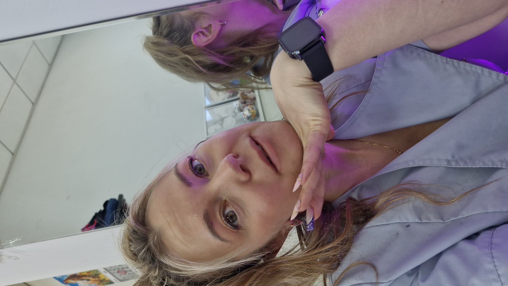 A woman is posing for a selfie in front of a mirror. She is wearing a light blue collared shirt. A black smartwatch is visible on her left wrist. Her hair is light brown with blonde highlights. She has long, manicured nails. The reflection in the mirror shows a portion of her face and hair, and a section of a white wall. The location is unknown and cannot be determined from this image.