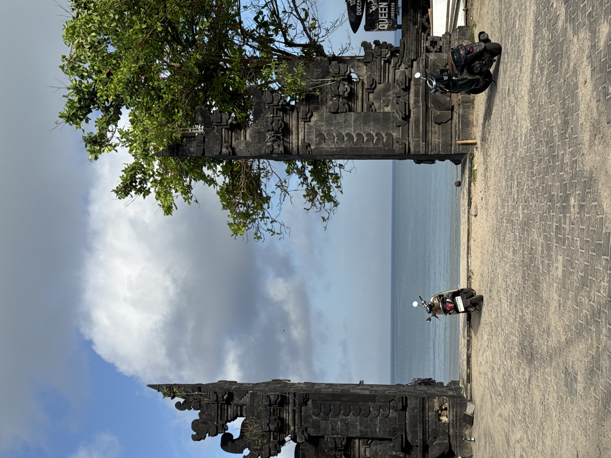 The image captures a scene from Jimbaran, Indonesia. A traditional stone gate frames a view of the sea and sky. The gate is intricately carved and weathered, with a tree growing beside it, partially obscuring the sky. The sky is cloudy, with a mix of blue sky visible in the lower left of the frame.
On the right side of the frame, two parked motorcycles are visible. The ground is paved with bricks or stones.