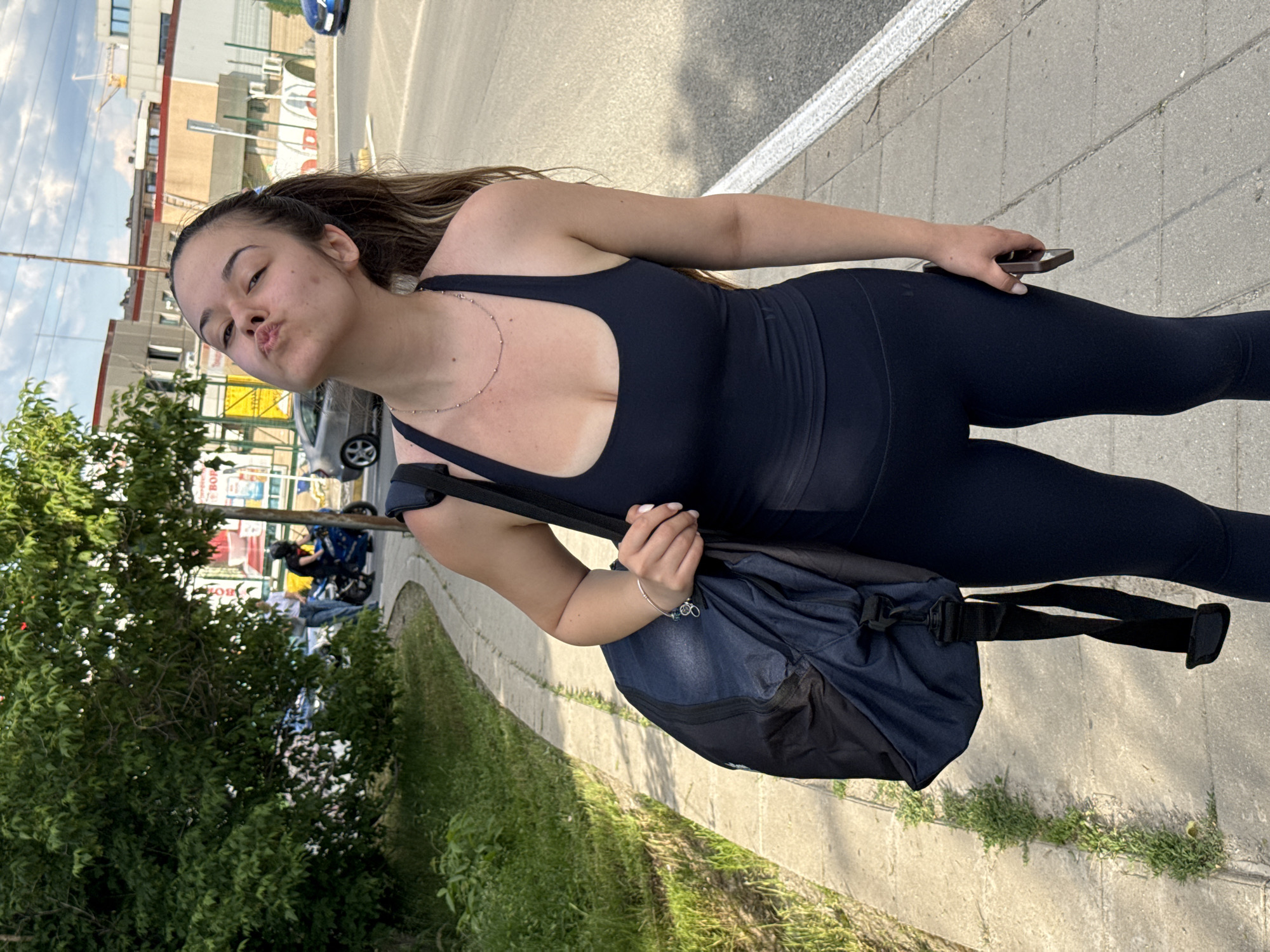 A young woman stands on a sidewalk in Sofia, Bulgaria, near a city street. She wears a dark-colored athletic outfit and carries a dark blue duffel bag over her shoulder. She holds a phone in one hand and poses with puckered lips. The background includes a street with parked vehicles, trees, and buildings, typical of an urban environment in Sofia.