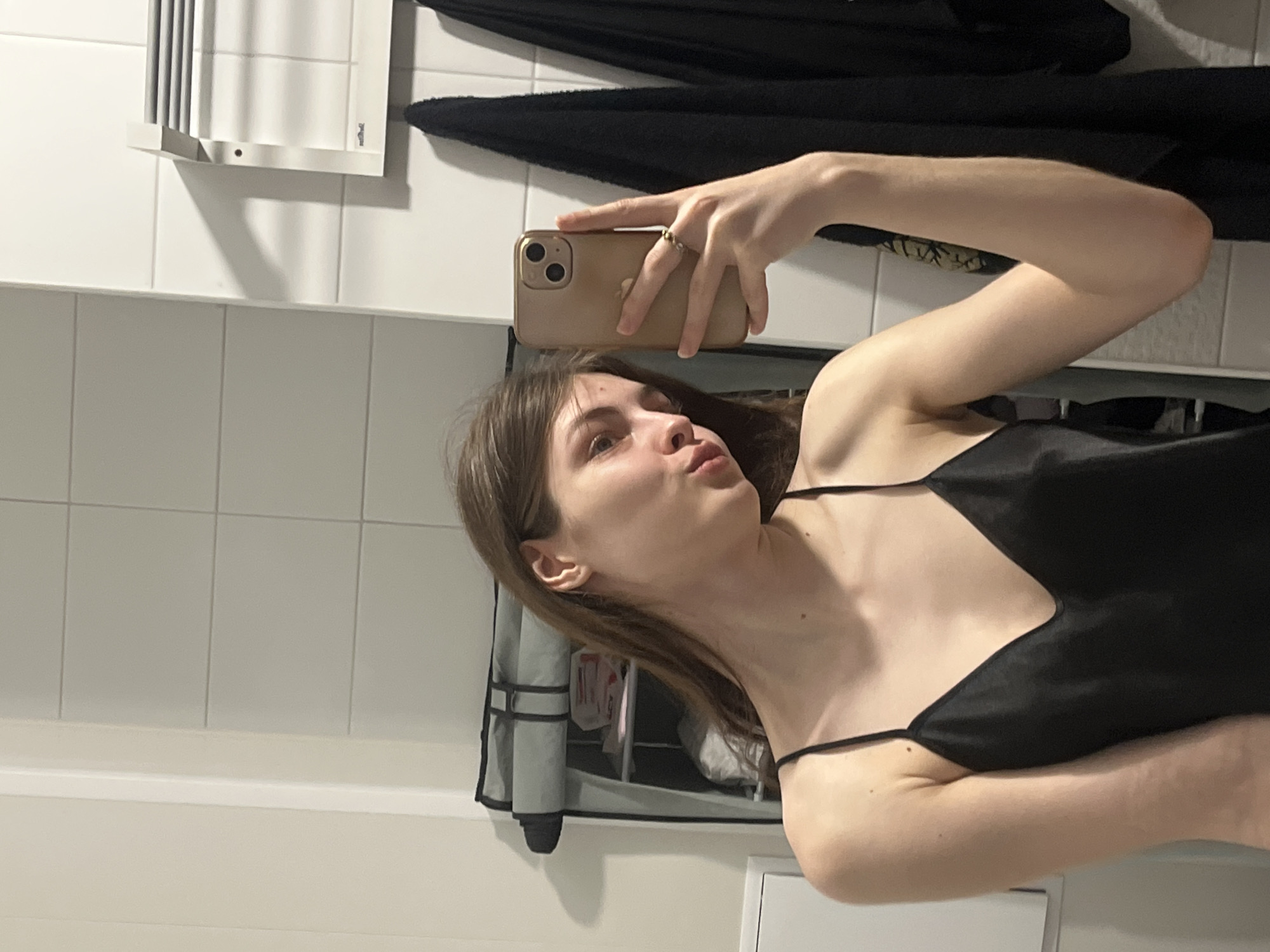 A young woman is taking a selfie in a bathroom. She is holding a gold-colored iPhone and posing with her lips pursed. She is wearing a black spaghetti-strap top. The bathroom has white tiled walls and includes a towel rack and hanging black towels. A partially visible cabinet is also present. The location in the city of Saint Petersburg, Russia, is not discernible from the provided visual information.