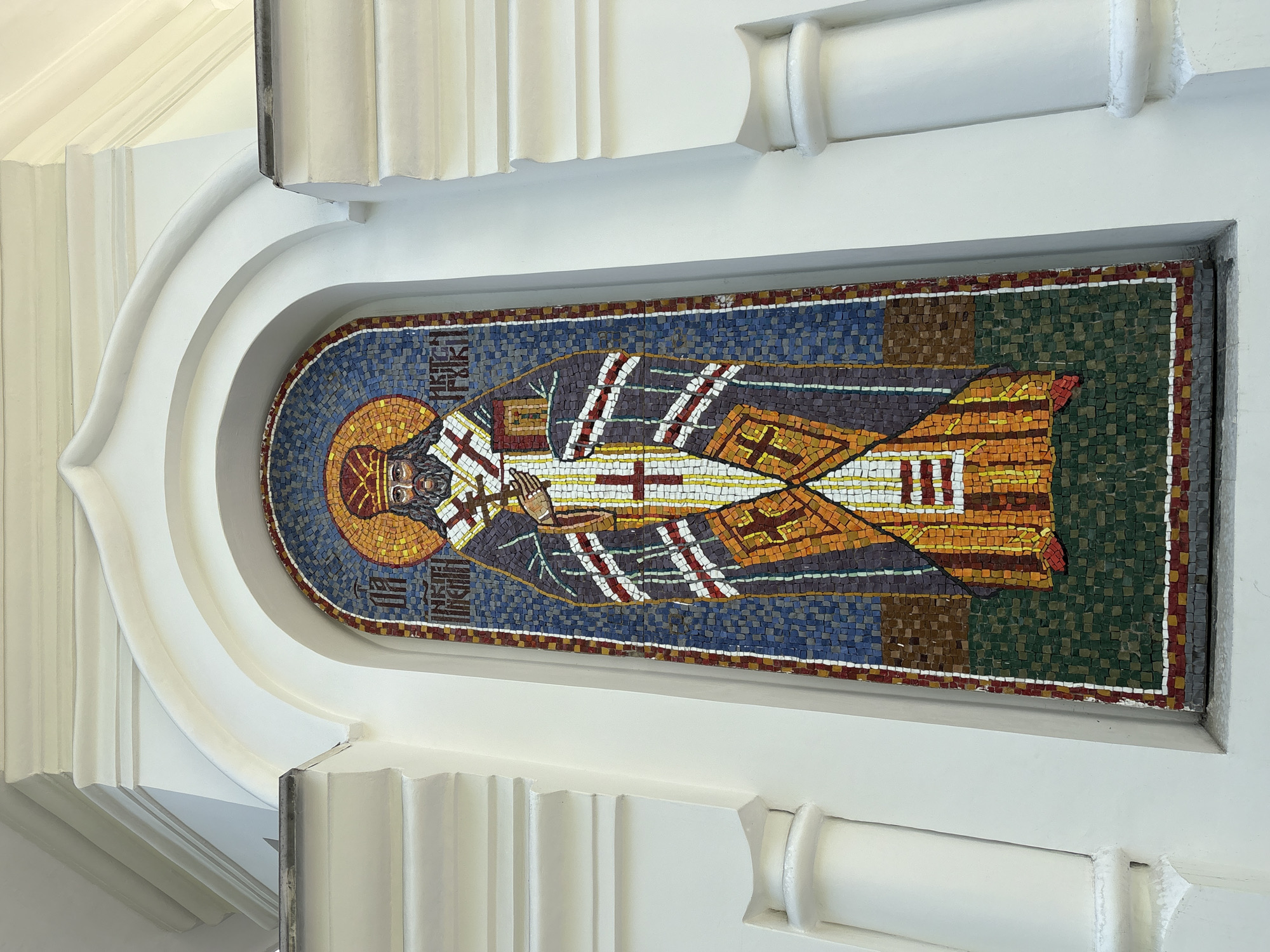 Here's a description of the image:
The image depicts a mosaic of a religious figure, likely an Orthodox saint or bishop, set within an architectural niche. The mosaic is composed of small, colored tiles forming a figure with a halo, wearing ornate vestments with crosses. The background is a mosaic of blue and green hues. The architectural niche is white with rounded arch and decorative fluted details on either side. The location is likely Birobidzhan, Russia, based on the prevalence of Orthodox iconography in the region.